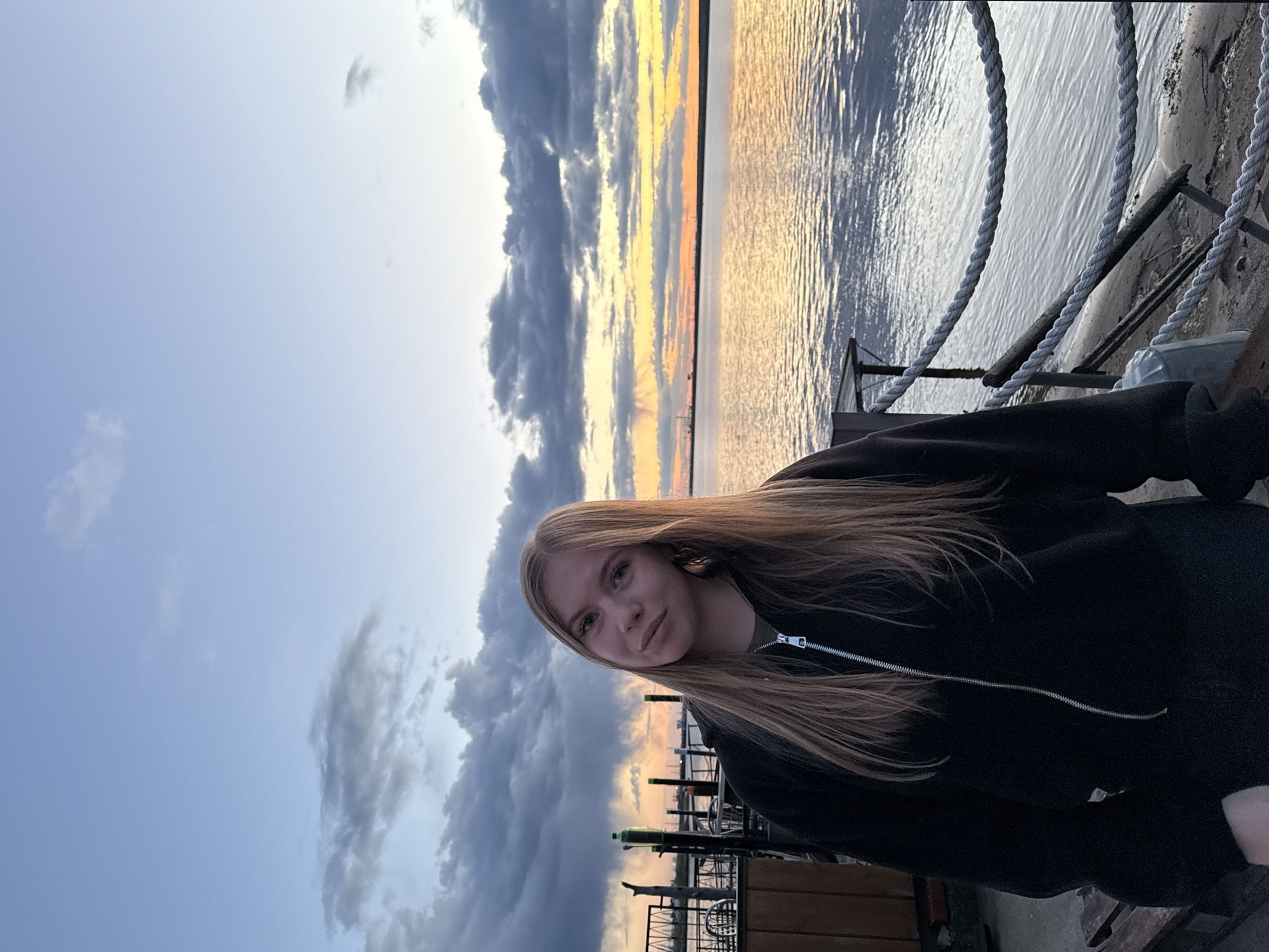 A young woman stands on what appears to be a pier or dock, likely at the embankment or riverfront of Chaikovsky, Russia, as the location features a body of water reflecting the colors of a sunset. The sky above is partially covered with clouds. The woman is wearing a black zip-up jacket. Ropes line the edge of the pier, and other structures are visible in the background.
