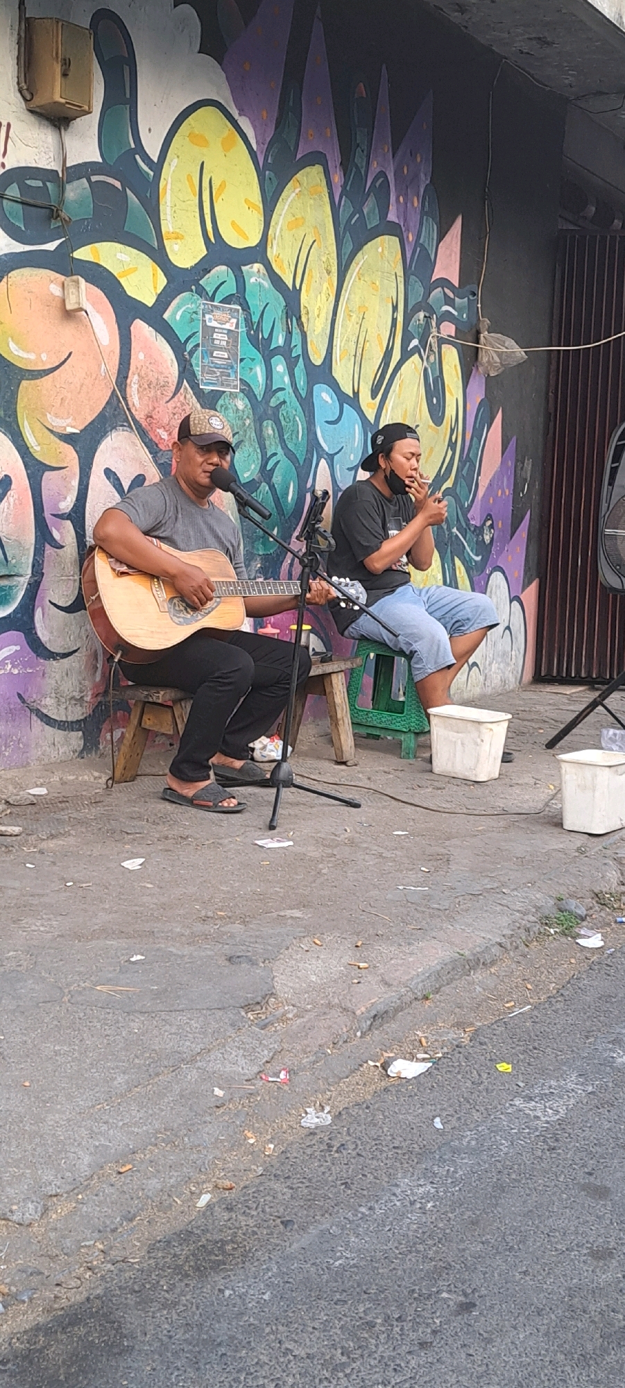 Two men are performing music on a sidewalk in front of a graffiti-covered wall in Petungasri, Indonesia. The man on the left is playing an acoustic guitar and singing into a microphone. He is wearing a gray t-shirt, black pants, and a baseball cap. The man on the right is sitting on a stool, smoking a cigarette, and wearing a black t-shirt, denim shorts, and a black baseball cap. The background features a colorful mural with abstract shapes. The ground is paved with asphalt and appears to be somewhat littered with cigarette butts and other debris. Two white buckets are placed on the sidewalk.