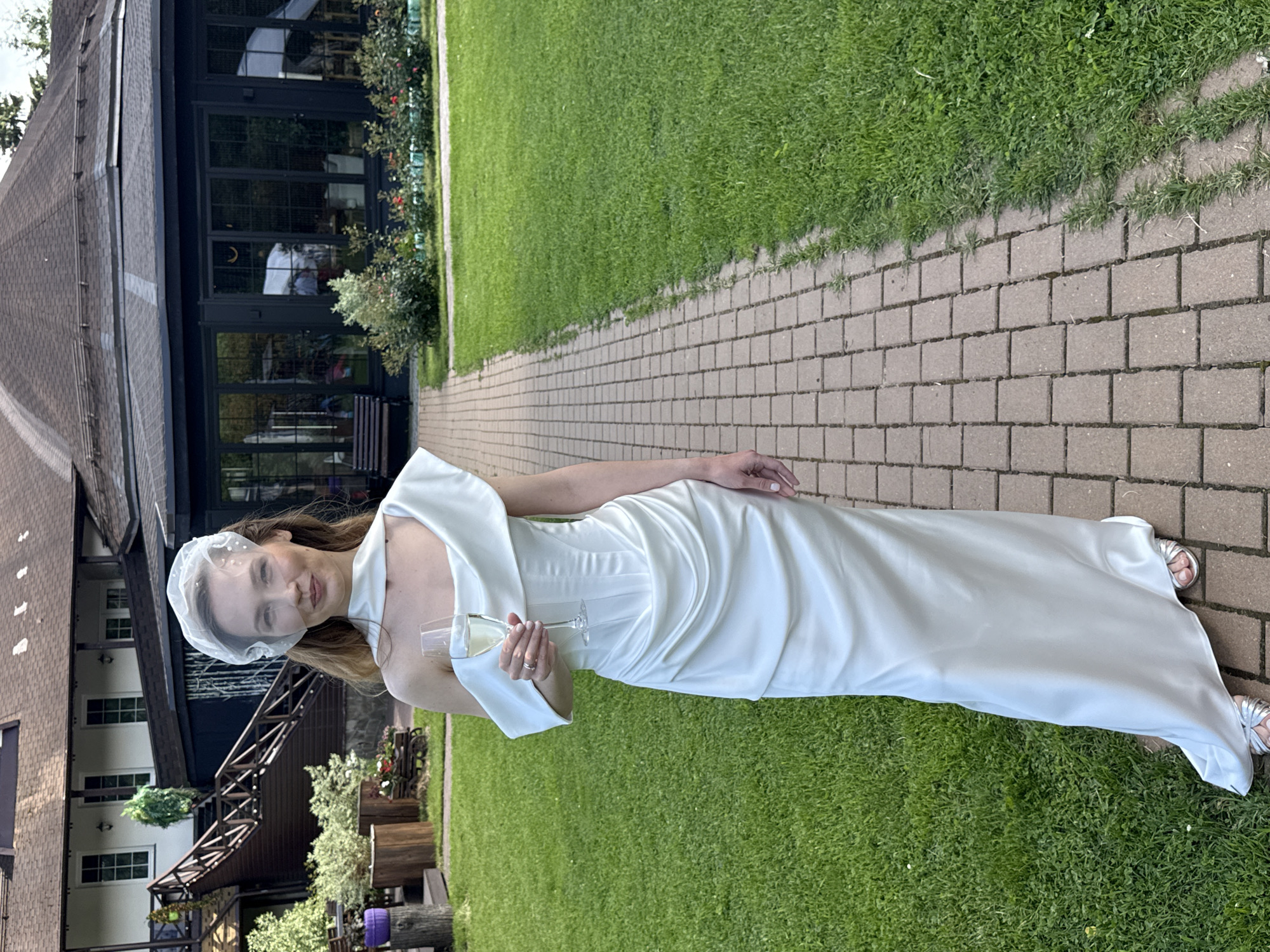 A woman in a white, floor-length dress and veil stands on a paved walkway next to a grassy area. She is holding a champagne flute in her right hand and looking towards the camera with a slight smile. Her dress has a draped neckline and appears to be made of a satin material. She is wearing silver sandals. In the background, there is a large building with a dark-colored roof and numerous windows. The building has a modern architectural style, with wooden accents and a multi-level structure. The surrounding area is well-maintained with green grass, bushes, and some wooden decorative elements. The lighting suggests it is daytime, possibly late afternoon or early evening, with a slightly overcast sky. The overall scene appears to be an outdoor event or gathering, possibly a wedding or a celebration, given the attire and the champagne. The location is identified as Khimki, Russia. There is no visible text in the image.