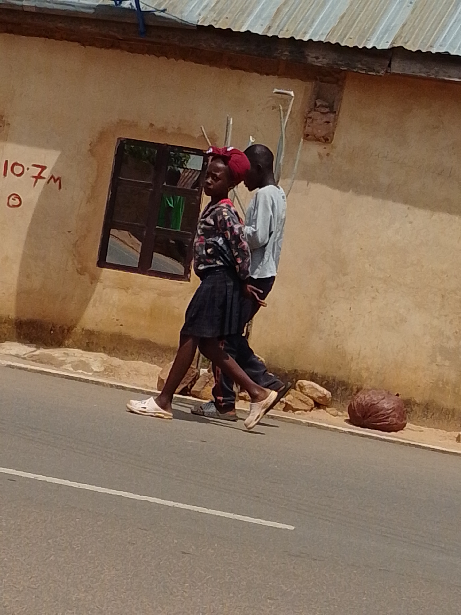 A full-shot, outdoor, eye-level photograph shows two children, a girl and a boy, walking on a road in front of a tan-colored, stucco building with a corrugated metal roof. The girl, on the left, is wearing a black skirt, a colorful patterned long-sleeved shirt, and white slides. She has red hair accessories. The boy, on the right, is wearing dark pants and a light blue long-sleeved shirt. The building has a dark-framed window with reflections of greenery inside. Red writing, reading "10.7M" and a circle, is visible on the wall to the left of the window. The road has a white line painted down the center. A dark, round object resembling a bundle of leaves or a large fruit is on the ground to the right of the children. The scene is illuminated by daylight.