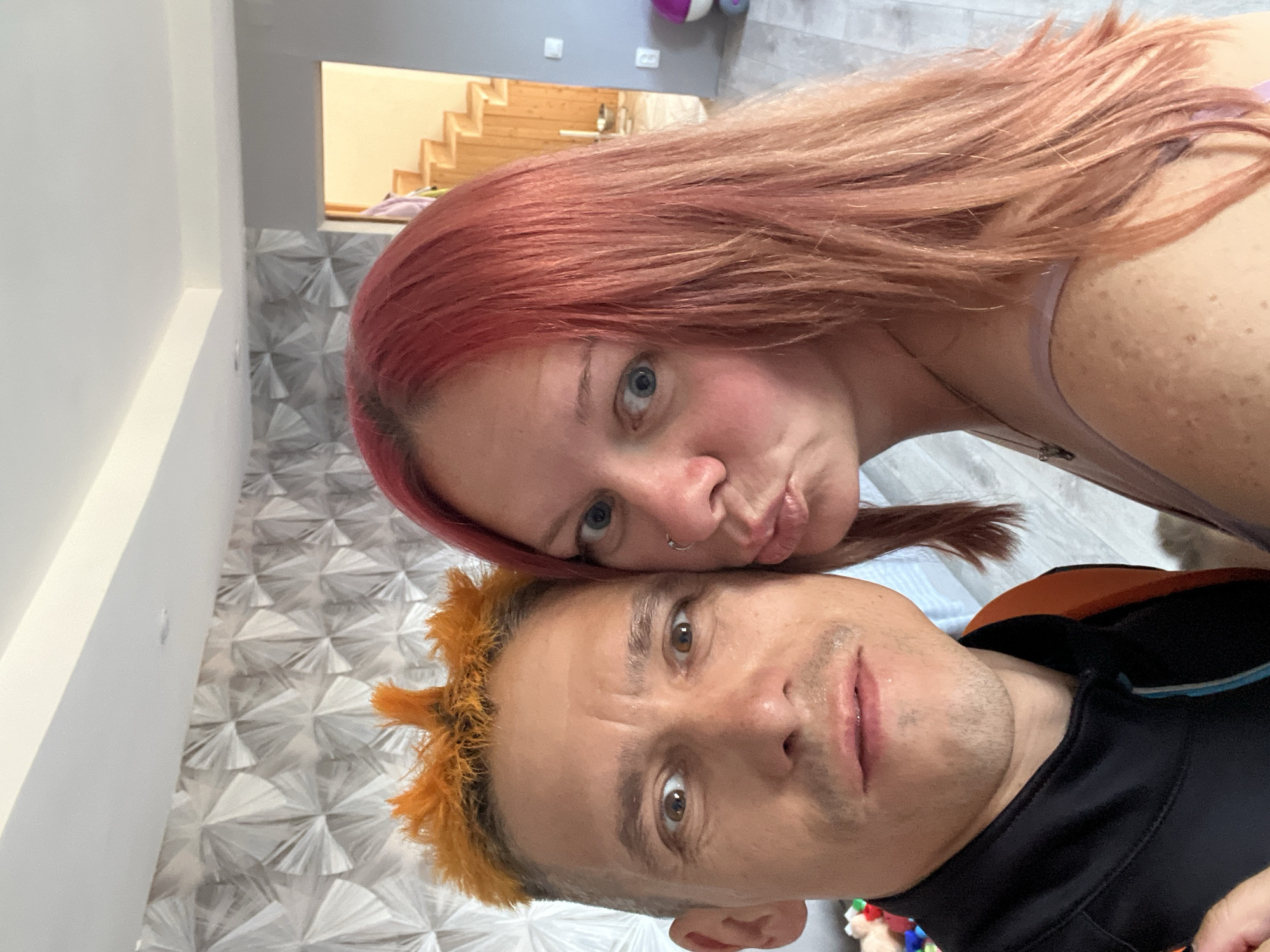 This is a selfie of a couple in what appears to be a home in Chaikovsky, Russia. The image is oriented vertically, with the couple's faces taking up most of the frame. The woman, on top, has reddish-pink hair and is looking towards the camera with a playful pout. She has blue eyes and a nose ring. The man, below her, has bright orange, spiky hair and is looking directly at the camera with wide eyes, his mouth slightly open. He has stubble on his chin. The background shows a modern interior with a patterned wall and what seems to be a staircase in the distance. There are no visible weather cues or specific time of day indicators, but the indoor setting suggests a casual moment. The overall emotion conveyed is one of lightheartedness and intimacy.
