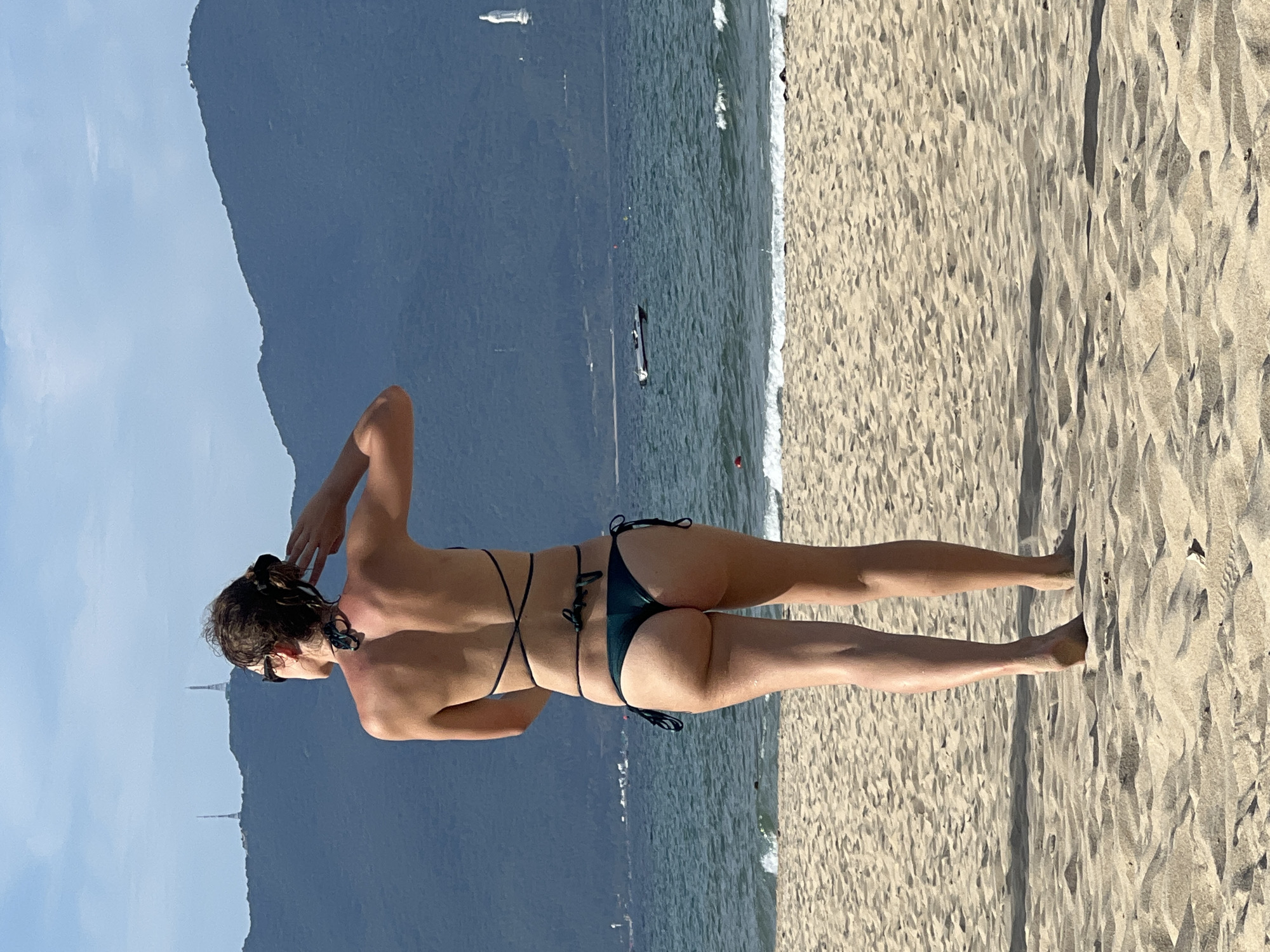 A woman in a dark green bikini stands on a sandy beach, facing away from the camera and towards the ocean. She has her hair pulled back and is touching the back of her head with her right hand. The ocean is calm with small waves, and in the distance, a chain of dark, tree-covered mountains rises under a bright blue sky with scattered clouds. A small boat is visible on the water near the mountains. The beach is composed of light-colored sand with subtle ripples and shadows, indicating a sunny day. The scene is consistent with the beaches of Đà Nẵng, Vietnam, known for their expansive sandy shores and scenic mountain backdrops.