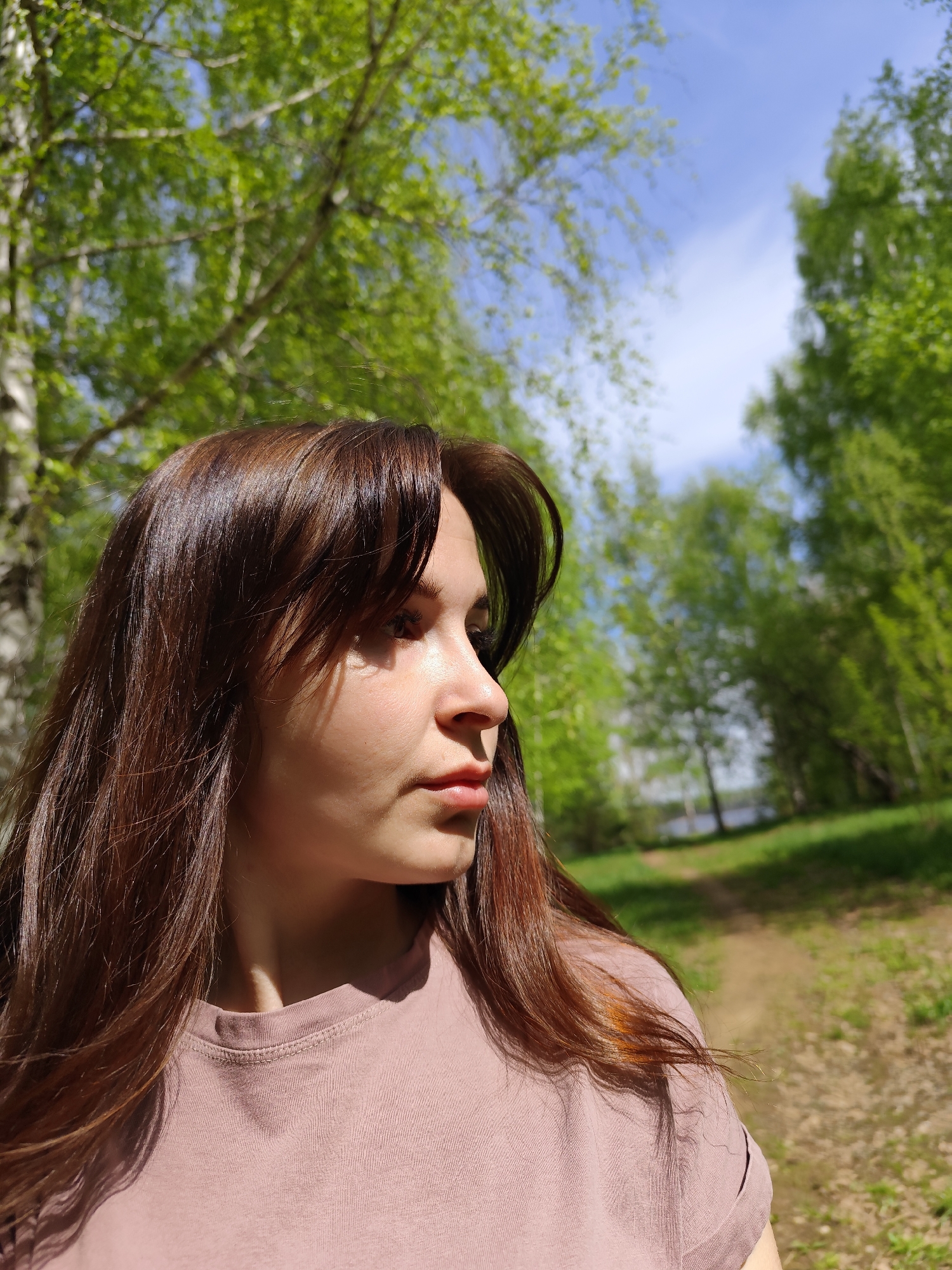 The photograph depicts a young woman with long, dark brown hair wearing a muted pink t-shirt. She is looking to the right, with a focus on something out of frame. The background consists of trees with vibrant green leaves, partially obscuring a blue sky with scattered clouds. A path is visible in the distance, flanked by more trees, leading toward a body of water.
The photograph was taken in the city of Chaikovsky, Russia.