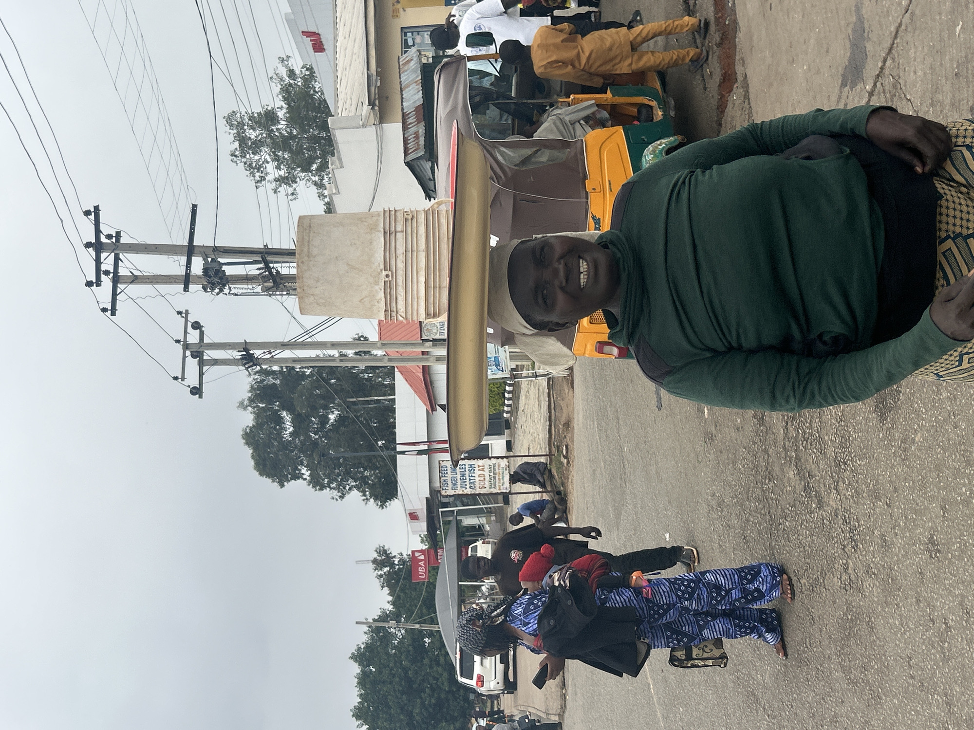The image shows a street scene in Jos, Nigeria. A man wearing a green shirt and patterned pants is in the foreground. He is smiling and appears to be carrying a large rectangular object on his head. Behind him is a yellow and green three-wheeled vehicle with people in it. In the background, there are buildings, trees, power lines, and other people walking on the street. A sign is visible on a building advertising fish feed and juvenile catfish. A woman in a patterned blue outfit is walking on the road carrying belongings.
