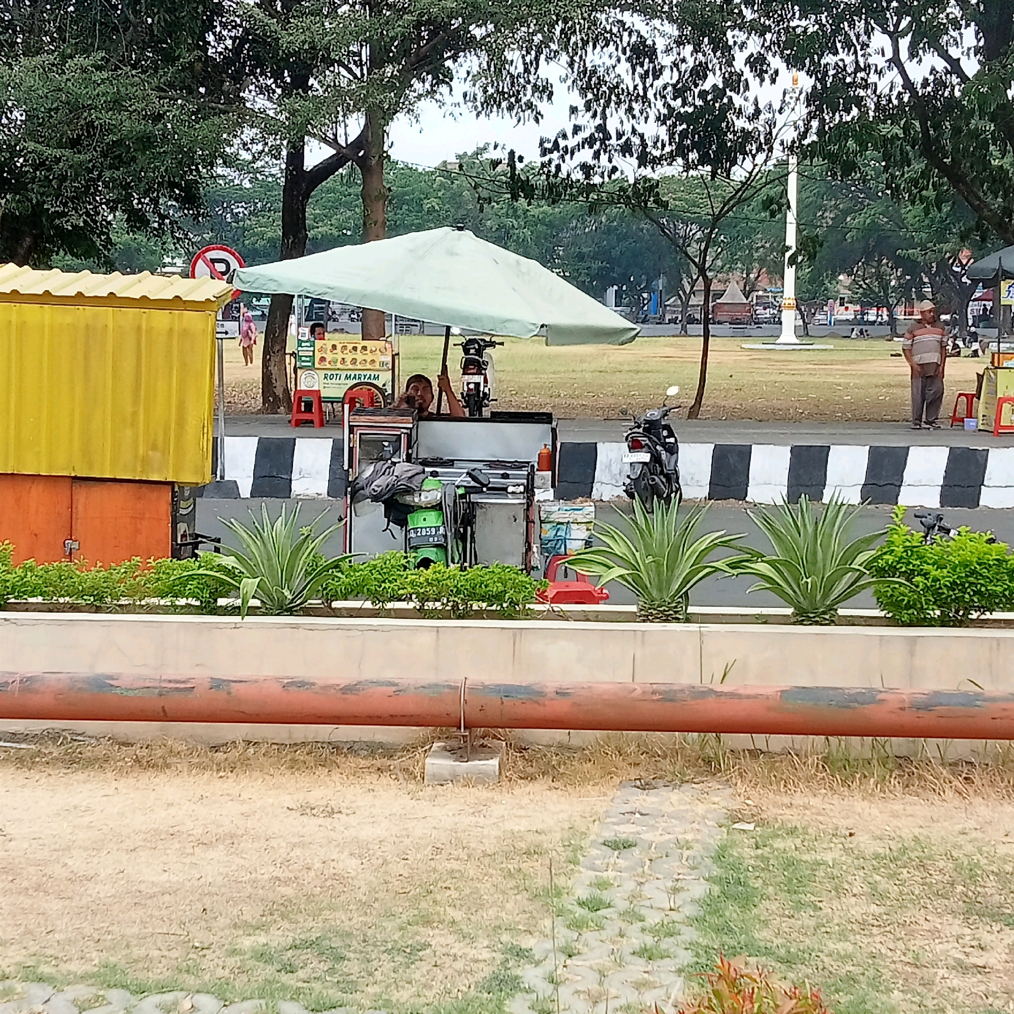 The image depicts an outdoor scene likely in the city of Karanganyar, Indonesia, possibly near a public park or recreational area. In the foreground, a large, rusty orange pipe runs horizontally across the frame, supported by concrete blocks. Beyond the pipe, a landscaped area features low green bushes and spiky succulent plants.
The mid-ground of the image reveals a street with a black and white striped pedestrian crossing. On the left side of the street, a bright yellow container-like structure is visible. Adjacent to this, a small food stall is set up under a light green umbrella. The stall has a cart displaying various food items and signage that reads "ROTI MARYAM." A person is partially visible behind the counter of the food stall, holding onto an umbrella pole. A motorcycle is parked behind the food stall.
Further back, across the street, is a large grassy field. In the distance, a white monument or flagpole stands prominently, with trees and buildings forming the background. Several people are scattered across the field, some sitting and others standing. Two men are visible near the right side of the frame. One man, wearing a striped shirt and cap, stands facing forward, while another man in a striped shirt and dark pants stands further back near another small stall with red stools. A pink-and-white striped signpost with a red circle and a diagonal line, indicating "No Parking," is also visible on the left side of the road. The overall atmosphere appears to be a casual, public gathering space, possibly during daytime.