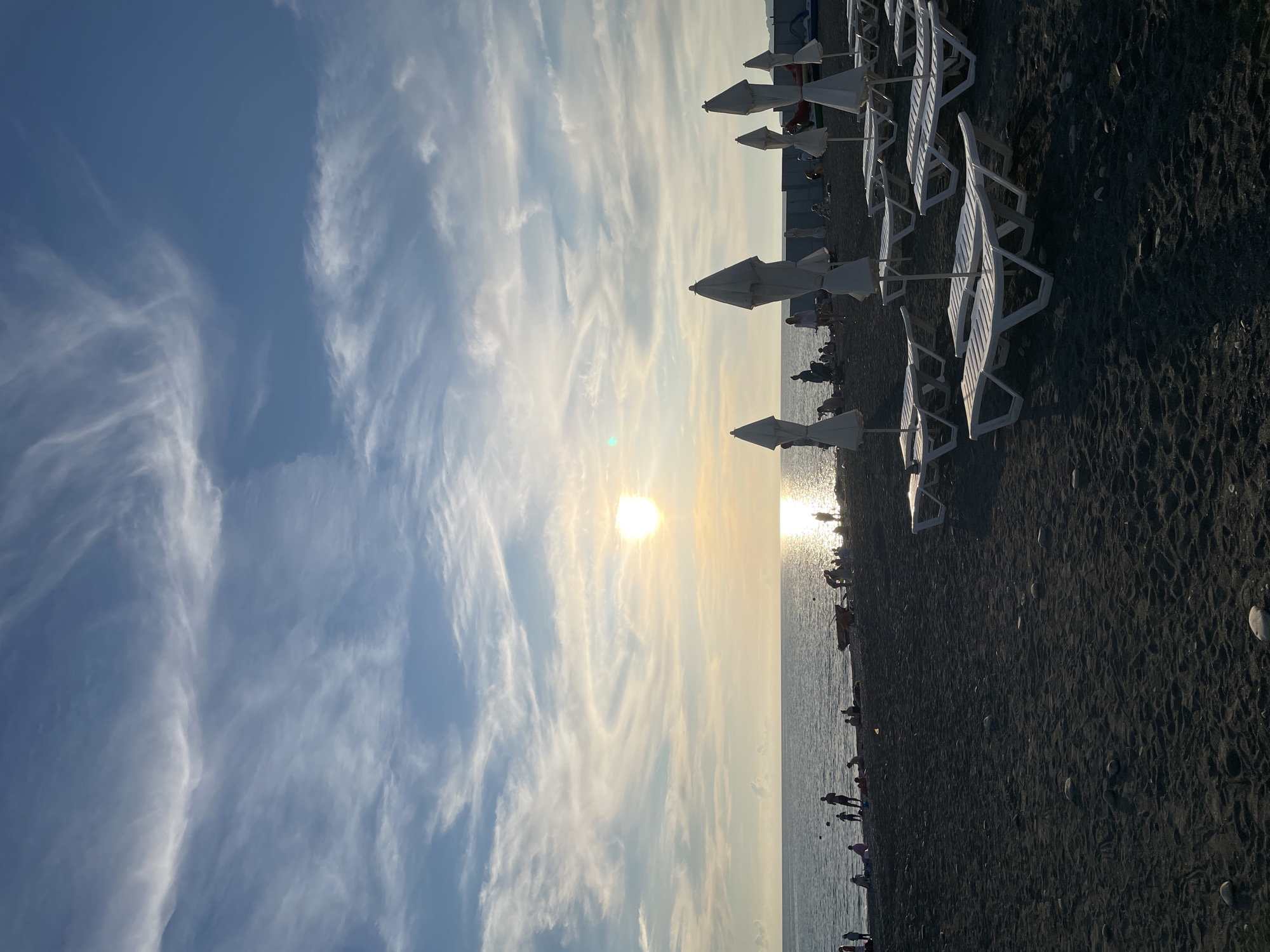 The image depicts a sunset scene on a black pebble beach, likely in Sochi, Russia. The sun is low on the horizon, casting a bright glare on the calm sea and illuminating the sky with wispy white clouds. In the foreground, rows of empty white plastic lounge chairs and closed white umbrellas are arranged on the dark sand, suggesting a commercial beach. A few people are scattered along the shoreline, some sitting and others standing, with a visible group of individuals gathered near the water's edge. The overall atmosphere is peaceful, with the warm glow of the setting sun creating a serene ambiance.