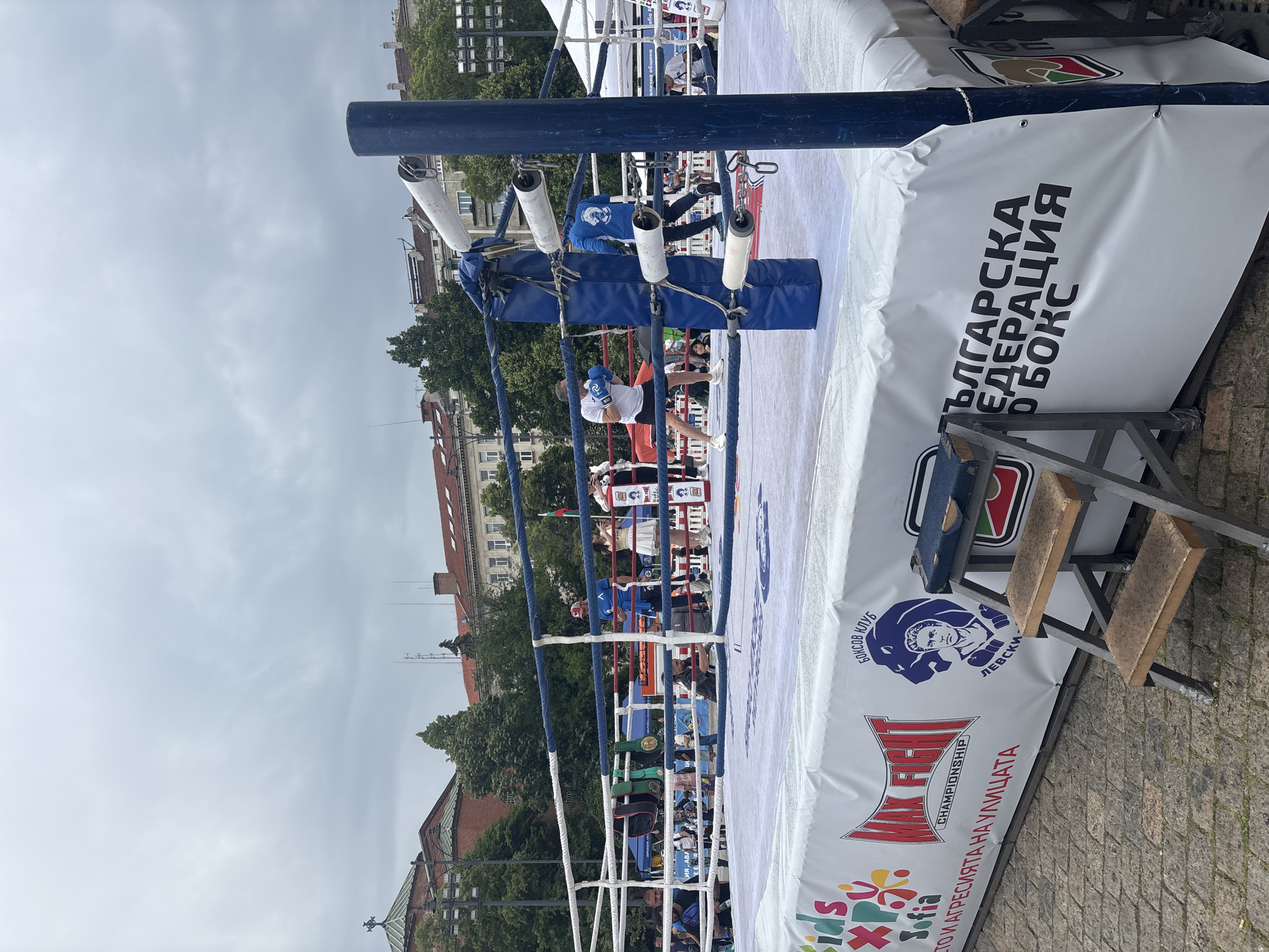 Here's a detailed description of the image:
**Scene:** An outdoor boxing ring setup in the city of Sofia, Bulgaria. The ring is the focal point. The backdrop consists of buildings, trees, and a cloudy sky.
**Boxing Ring Details:** The ring structure is constructed of metal posts and ropes, covered in white protective material. The ropes are padded with cylindrical padding in blue and white. The side of the ring is adorned with advertising banners and logos. Prominently displayed are the words "БЪЛГАРСКА ФЕДЕРАЦИЯ БОКС" (Bulgarian Boxing Federation). There's also a logo for "БОКСОВ КЛУБ ЛЕВСКИ" (Boxing Club Levski) and a logo that reads "FIGHT CHAMPIONSHIP". At the bottom, it says, "СТО И АГРЕСИЯТА НА УЛИЦАТА" (Stop the Aggression in the Street). The word “Kids” is prominently displayed at the bottom left. There is a metal step ladder resting against the side of the ring.
**People:** There are boxers in white clothing, within the boxing ring. There are also people wearing blue standing ringside.
**Environment:** The image appears to be taken at ground level. The background includes multi-story buildings. The sky is overcast. The ground around the ring appears to be paved.