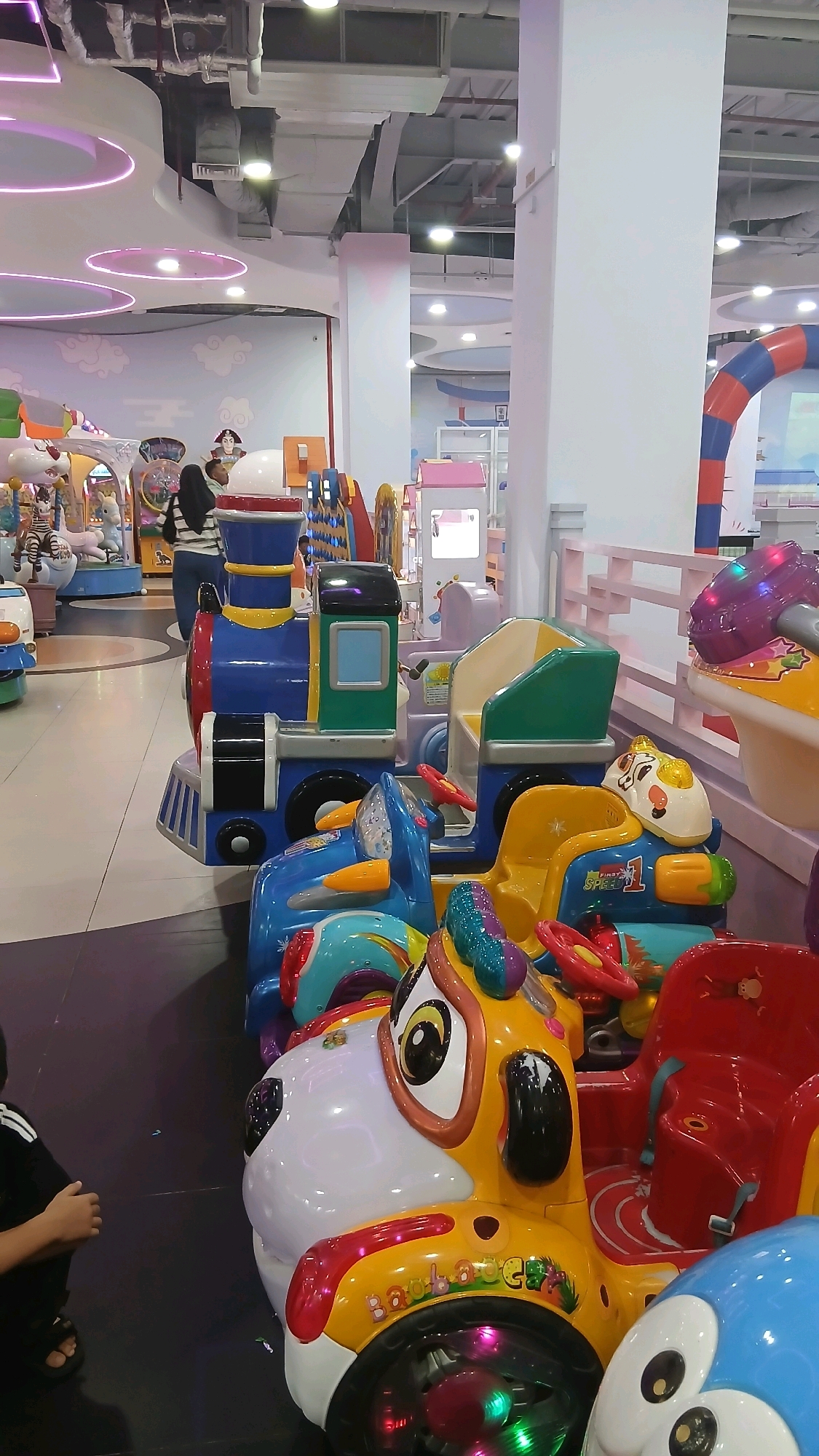The image shows a row of colorful children's amusement rides, resembling a train and various animal-themed cars, situated inside an indoor entertainment facility. The rides are brightly lit with LED lights, and some have cartoonish faces and vibrant paint jobs. In the background, there are other play structures, including what appears to be a large archway and soft play equipment. Two individuals, one wearing a hijab, are visible in the mid-ground, looking at the rides. The floor is tiled, and the ceiling features modern lighting and ventilation systems. The overall impression is a lively and child-friendly environment, typical of a family entertainment center or arcade. This scene is located at Asia Mega Mas Mall in the City of Binjai, Indonesia.