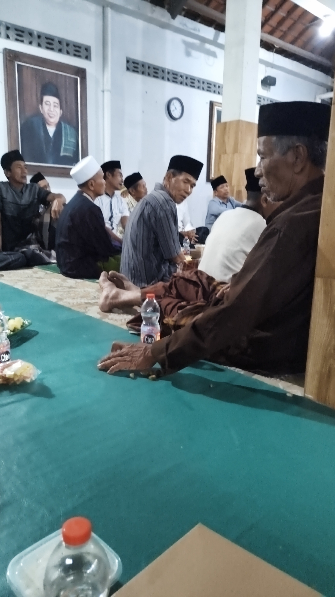 In Dalegan, Indonesia, a group of men are seated on a teal-colored mat inside a building with a tiled roof and white walls. In the background, a framed portrait of an older man in traditional attire hangs on the wall. To the right of the portrait, a round clock is visible. Several men, most wearing peci hats, are gathered in conversation or observation. One man in the foreground, wearing a brown shirt and peci, is seated facing right, with his hands resting on his lap, appearing to interact with small items on the mat. Another man to his left, wearing a striped shirt and peci, is looking to his left. A bottle of "Cleo" mineral water is placed on the mat in front of the man in the brown shirt. The environment appears to be a communal gathering space.