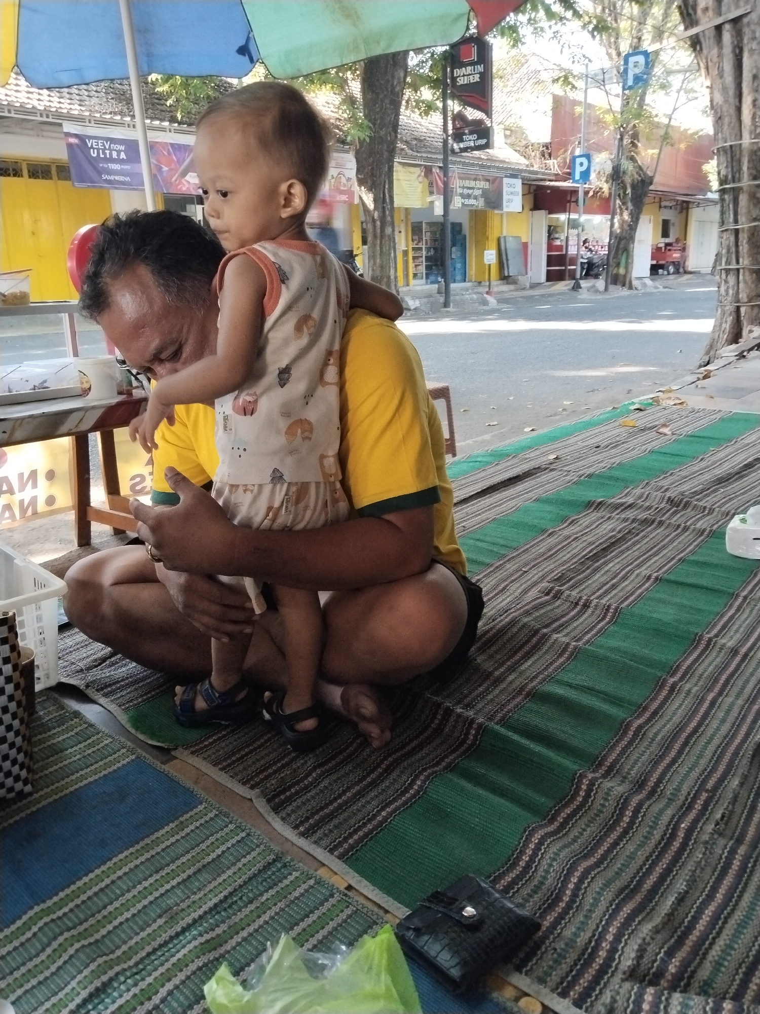 A man in a yellow t-shirt with a child on his shoulders is sitting on a striped mat on the ground. The child is wearing a patterned sleeveless shirt and shorts. The man is looking down at the child. In the background, there are shops with signage in Indonesian. One sign reads "DIARUM SUPER". There are trees and a road visible. Parking signs with a "P" are present. The scene appears to be outdoors on a sunny day.