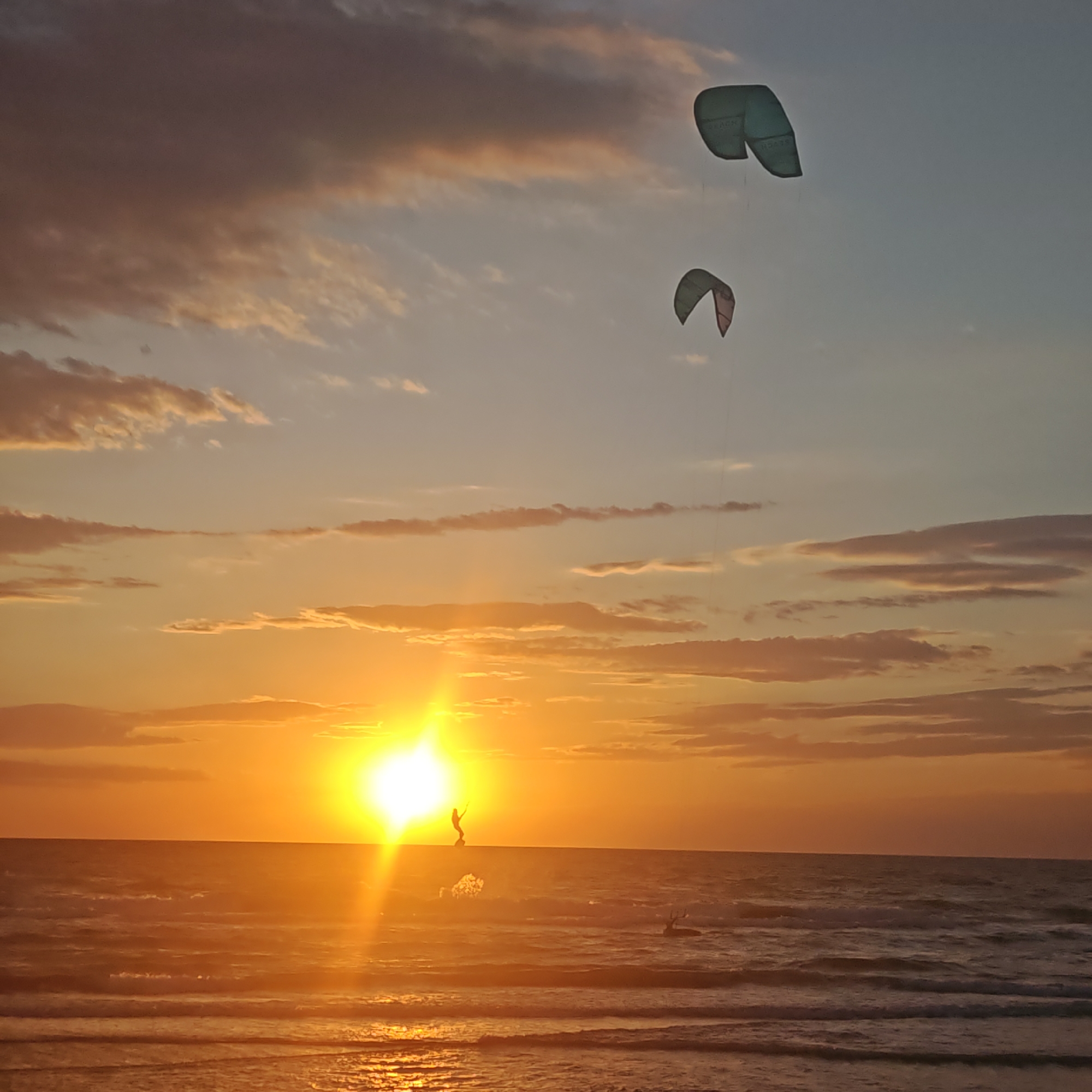 The image depicts a sunset over the sea in городское поселение Щелкино, Ukraine. The sun is low on the horizon, casting a bright glow and long rays across the water. There are two kites in the air, tethered to kiteboarders in the sea. One kiteboarder is near the horizon in direct line with the sun, appearing silhouetted. The sea has small waves, and the sky is filled with clouds tinged with orange and yellow hues from the setting sun.