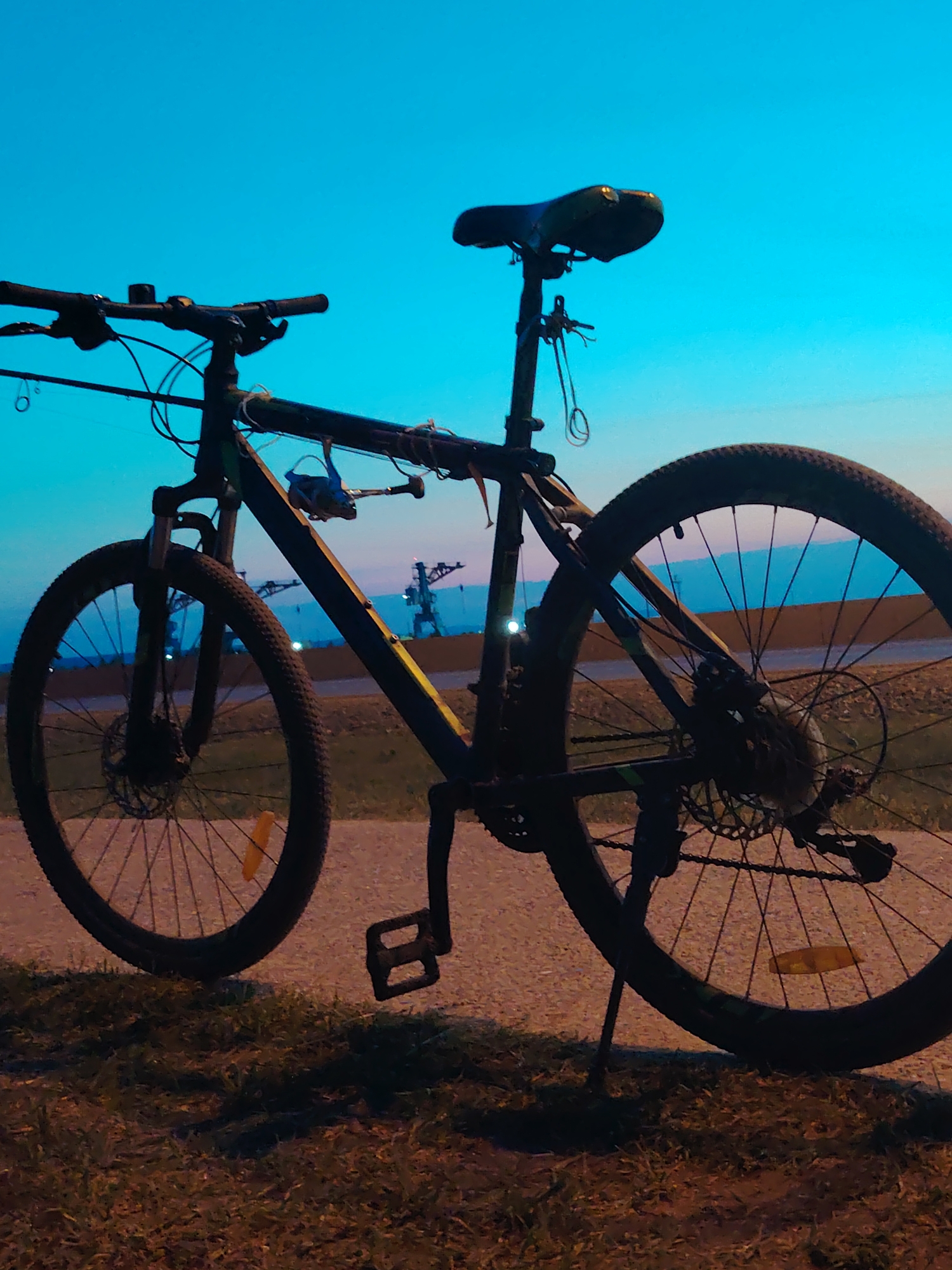 A bicycle is parked on a paved surface next to a grassy area. A fishing rod and reel are mounted on the bicycle's frame. In the background are structures including cranes. This picture was taken in the city of Chaikovsky, Russia. The sky is blue. It is dusk.