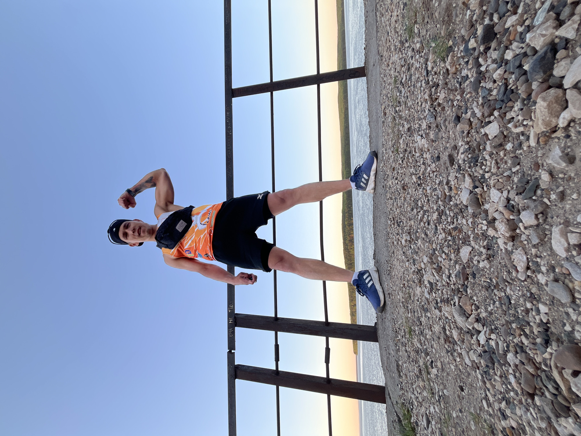 A man stands at a metal railing on a rocky embankment in Chaikovsky, Russia. He wears a black cap, an orange and white patterned tank top, black shorts, and blue sneakers. He also has a small black bag strapped across his chest. His arms are flexed, showing a tattoo on his left arm. The railing is made of dark metal and has a simple design. Behind the railing, there is water and a thin strip of land in the distance, with the sky visible above. The embankment is covered in rocks and pebbles. The sky is light blue near the top of the image, fading to a yellow-orange near the horizon.
