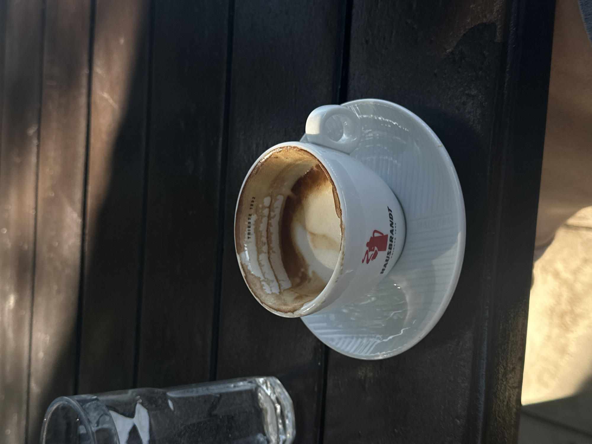 An overhead shot shows a dark wooden table with a half-empty cup of coffee and a glass of water. The coffee cup is white and sits on a white saucer. The cup has a logo that reads "HAUSBRANDT TRIESTE 1892." The coffee inside is mostly gone, with only a small amount of foam and brown liquid remaining at the bottom. The glass of water is to the left of the coffee cup and is partially filled. The table has a weathered appearance with visible grain and shadows cast by sunlight. The scene appears to be outdoors, possibly on a patio or balcony.