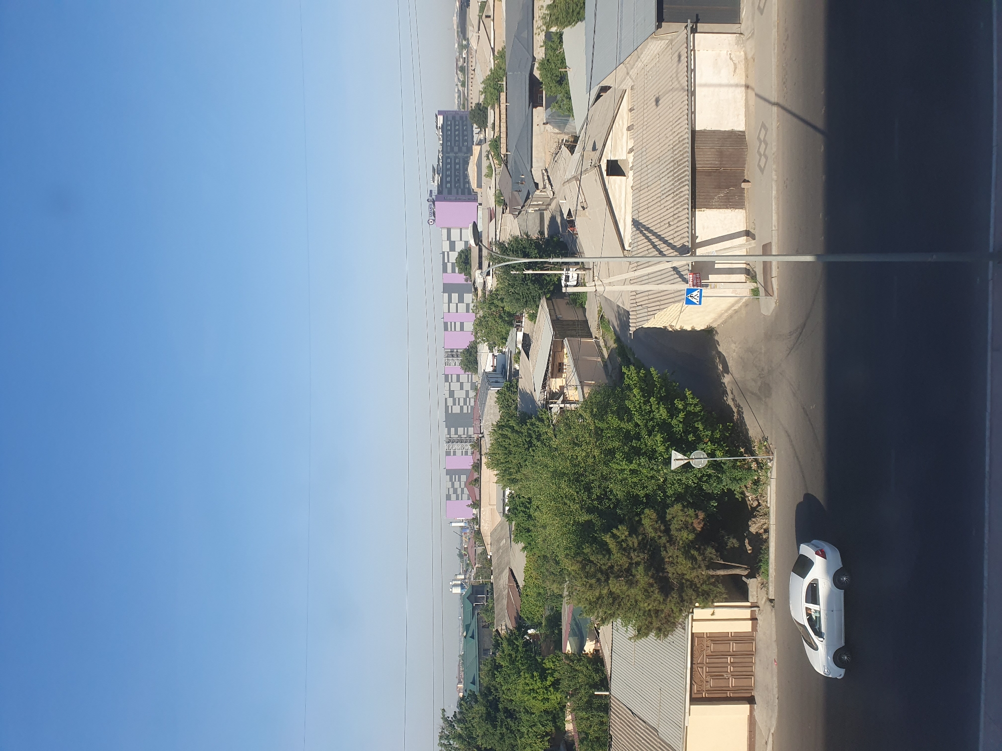 The image shows a wide view of a street in Tashkent, Uzbekistan, under a clear blue sky. In the foreground, a white sedan is driving on the road. The street is lined with single-story houses with flat, corrugated roofs and beige-colored walls. Some houses have large, dark-colored doors. Trees with lush green foliage are scattered throughout the scene, interspersed between the buildings. In the background, a large, modern building complex with purple and gray sections is visible, bearing a logo that appears to say "UZUM." Streetlights and power lines are present. The overall scene depicts a typical urban residential area in Tashkent.