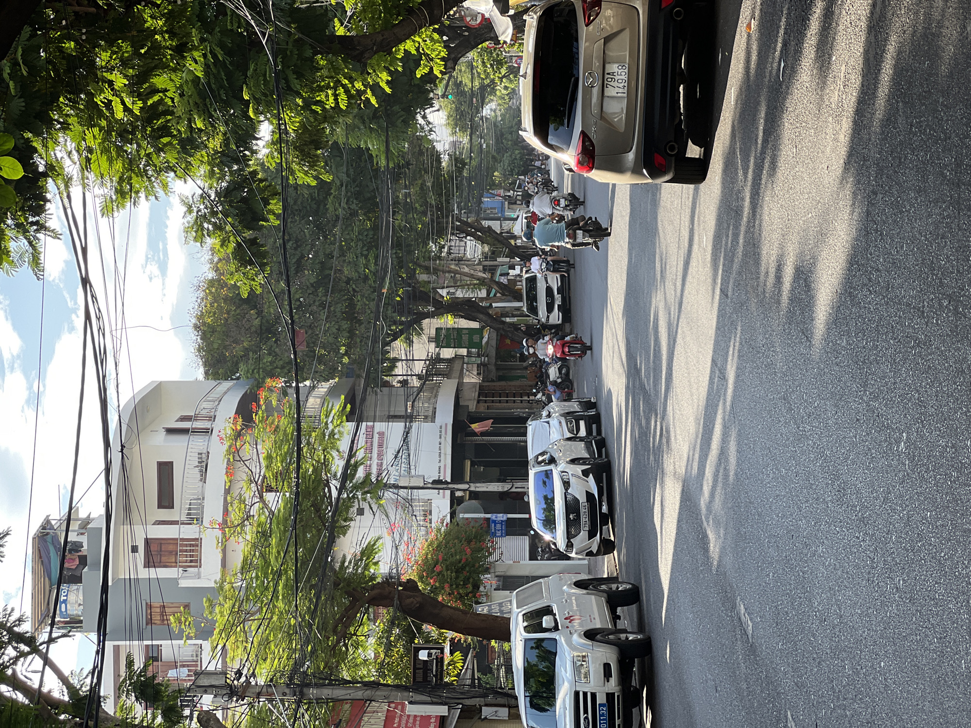 The image depicts a street scene in Nha Trang, Vietnam, with a focus on the road and surrounding buildings. In the foreground, a beige SUV, identified by its license plate "79A 149.58," is parked on the right side of the road, facing away from the camera. To the left, a white pickup truck with license plate "011.32" is also parked.
The street is paved with asphalt and shows some signs of wear. Several other vehicles, including white cars and motorcycles, are visible further down the street, indicating moderate traffic. Pedestrians are present, primarily on motorcycles. Buildings line both sides of the street, featuring a mix of architectural styles. Some buildings have multiple stories with balconies, while others appear to be commercial establishments with storefronts. Overhead, a complex network of electrical wires crisscrosses the scene. Green trees with vibrant foliage and some flowering trees add a natural element to the urban environment. The sky is partly cloudy with patches of blue visible. The lighting suggests it is daytime, with sunlight casting shadows across the road.