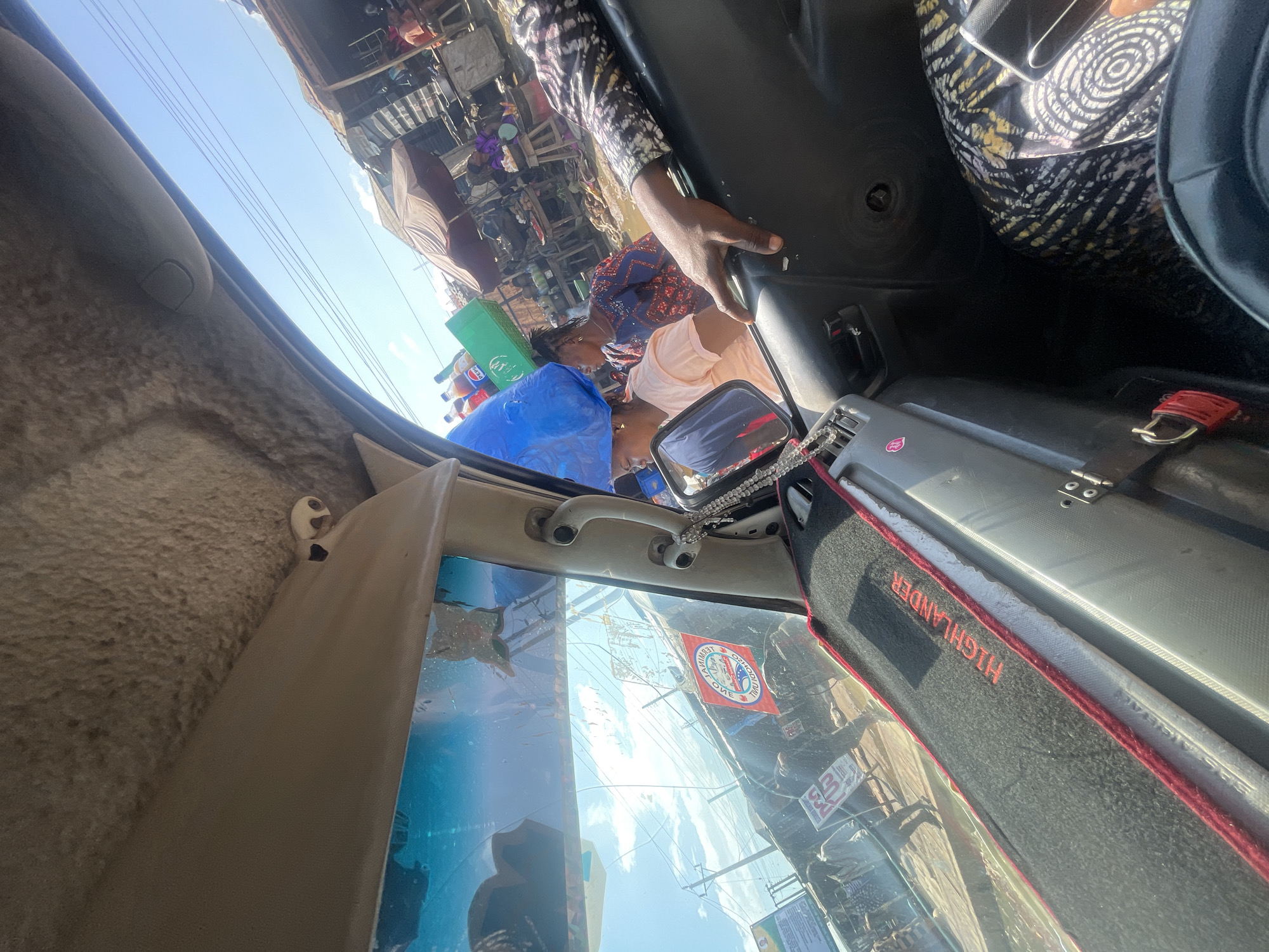 This media file captures a dynamic scene from inside a vehicle, likely a bus or van, looking out. The perspective is from the passenger side, facing forward and slightly upwards. The primary subjects are several individuals, with a focus on two women in the front of the vehicle. One woman, wearing a blue and red patterned top, is looking down, while another woman, in a light pink shirt, is looking towards the side. Their expressions are not clearly discernible.
The scene outside is a bustling market or roadside setting in Mowe, Nigeria. It appears to be daytime, with bright sunlight indicating a clear sky. The environment is filled with stalls, structures, and a crowd of people, suggesting a busy commercial area. There are various objects visible, including a large blue plastic bag, a green crate, and several bottles of what appear to be beverages, possibly soda. The vehicle's interior is also visible, with a textured tan panel on the left and a black dashboard area on the right. A grey suitcase with red trim is placed on the right side of the vehicle's interior.
A notable detail is a red flag with text that reads "CHAMPION" and "MUMBAI" with a circular emblem. There's also a red and black mat on the dashboard with the word "HIGHLANDER" in red lettering. The overall impression is one of vibrant activity and movement, characteristic of a busy market or transportation hub in Nigeria. The image appears to be taken during the day, with strong sunlight casting shadows and illuminating the scene.