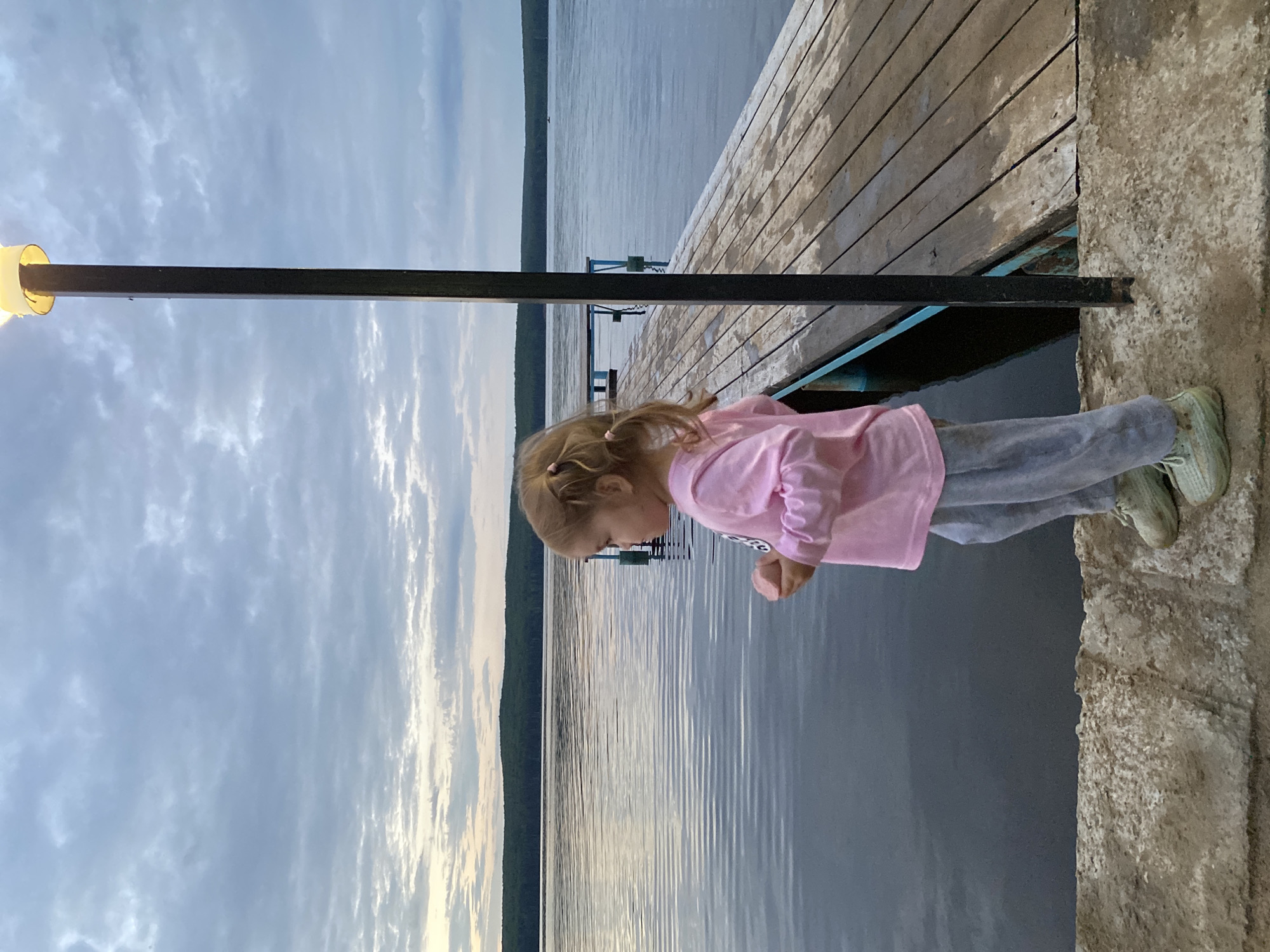 A young girl with blonde pigtails, wearing a pink long-sleeved shirt and gray sweatpants, stands on a wooden pier. She is holding a small, light-colored object in her right hand. The pier extends into a calm body of water, with a distant shoreline covered in trees under a cloudy sky. A black lamppost with a lit yellow fixture is positioned to the right of the girl. The scene appears to be a serene outdoor setting, possibly on the Kama River near Chaikovsky, Russia, given the landscape and pier structure.
