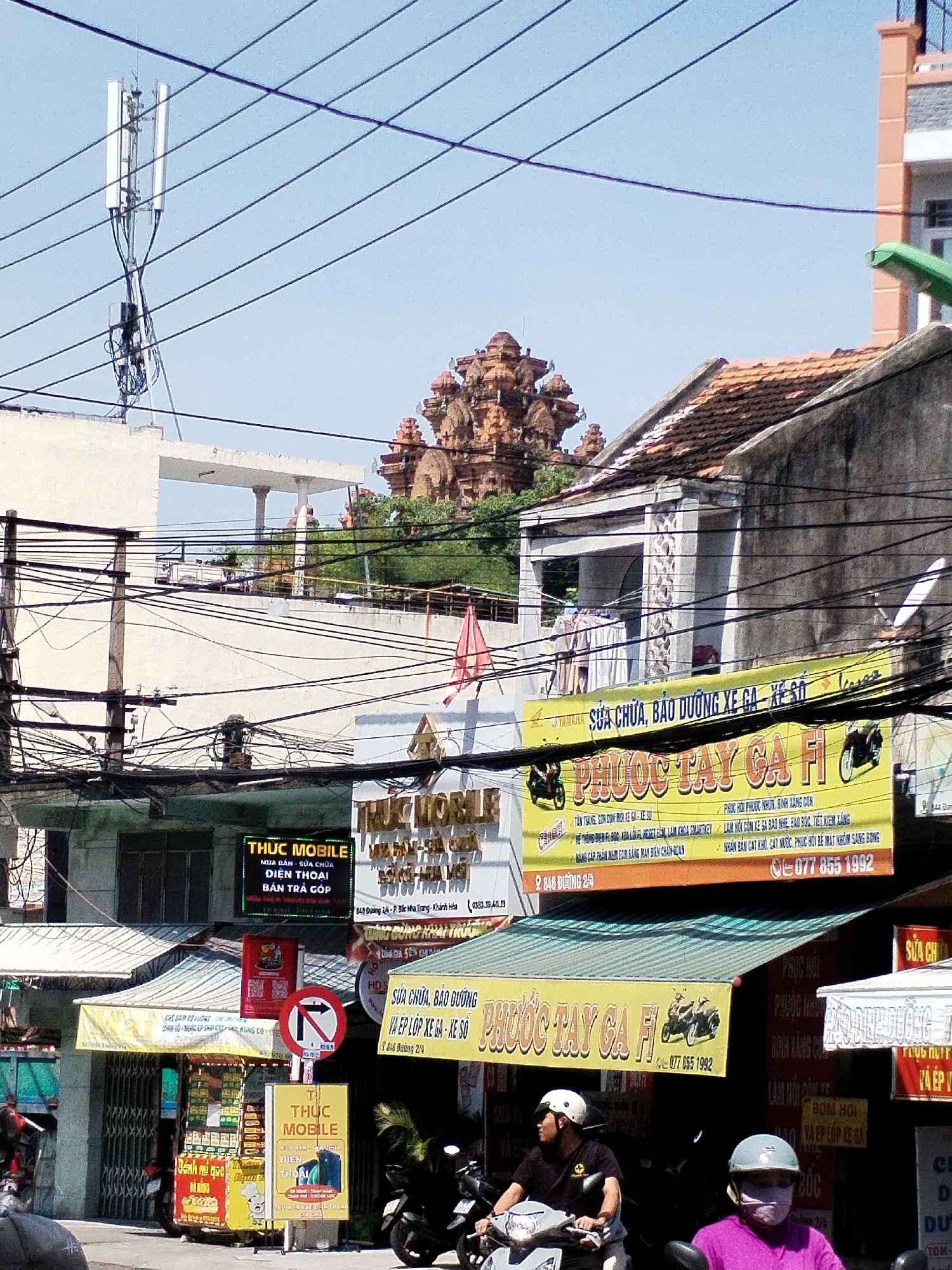 In the city of Bac Nha Trang Ward, Vietnam, a street scene unfolds under a clear sky, marked by a dense network of electrical wires crisscrossing the frame. A cellular tower stands prominently on the left, its multiple antennae reaching upwards. Below, a complex infrastructure of wires, poles, and various buildings suggests a bustling urban environment. The architecture includes a prominent structure with a red tile roof and decorative elements. Several signs advertise services like "THUC MOBILE" and "PHUOC TAY GA FI," indicating mobile phone sales, repairs, and motorcycle services. These signs are predominantly in yellow and black, with Vietnamese text. In the foreground, two individuals on motorcycles, wearing helmets, are visible, suggesting ongoing traffic or a pause in their journey. The overall atmosphere is one of a busy street in Vietnam, with a strong visual presence of communication infrastructure and commercial signage.