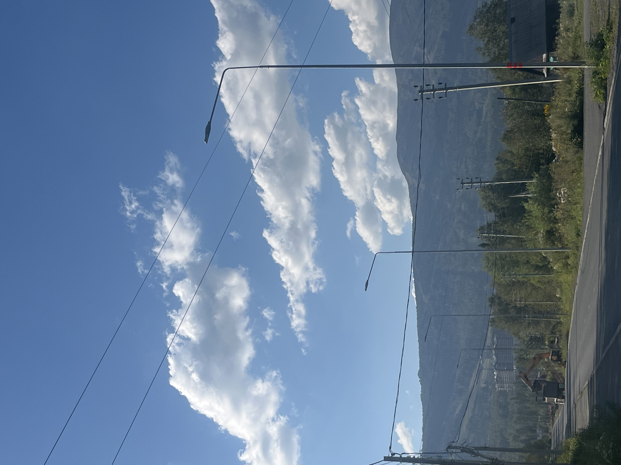 The image depicts a view along a paved road in the city of Sheregesh, Russia, under a bright blue sky with scattered white clouds. On the left side of the road, there are power lines supported by poles, and a construction site with an excavator and a dump truck is visible. In the distance, a large, multi-story building with a repeating architectural pattern stands amidst a dense forest. The road stretches forward, flanked by vegetation and trees. On the right side of the road, there is a dark-roofed building and more power poles. In the background, a gently sloping, tree-covered mountain range dominates the horizon. The overall scene suggests a developing area within a natural, mountainous landscape.