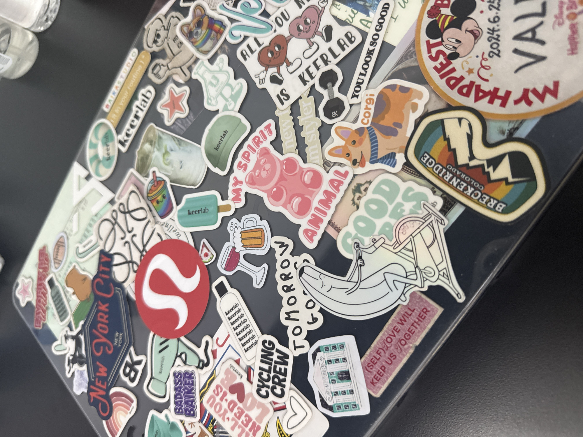 The image shows a laptop lid covered in stickers. The stickers feature a variety of designs, including logos, illustrations, and text. Prominent stickers include "New York City," a red omega symbol (likely a Lululemon logo), "Cycling Crew," "Breckenridge Colorado," a corgi, and "keerlab" repeated multiple times. The laptop is sitting on a dark surface. The location is unknown.