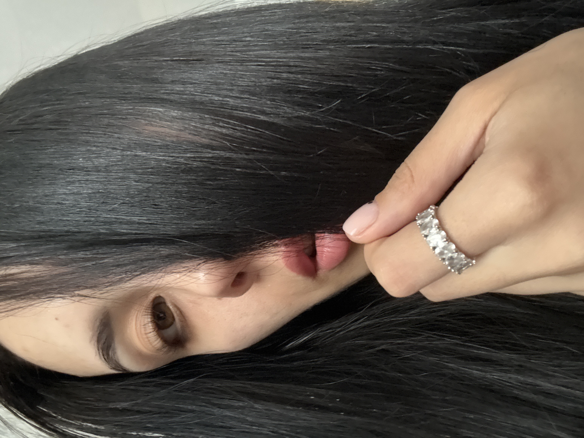 A close-up, portrait-style photograph features a person with long, dark hair partially covering their face. Their face is turned to the side, revealing one eye with long eyelashes, part of their nose, and lips. The skin appears to be fair. The person's hand, adorned with a silver ring set with rectangular stones, is holding a strand of hair near their lips. The background is a plain, light color. There is no information in the image to indicate the location is Votkinsk, Russia.