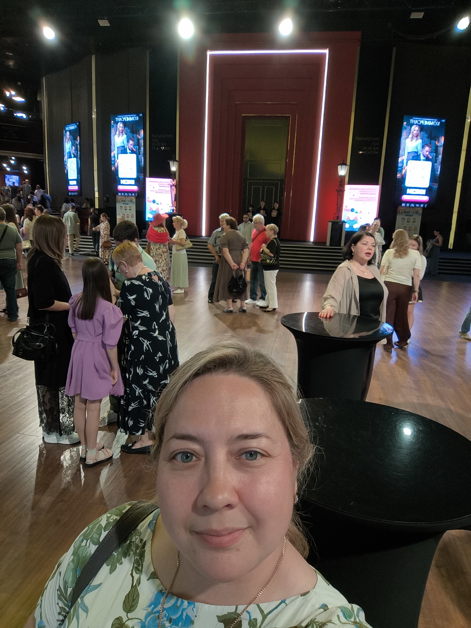 A woman with blonde hair and blue eyes is taking a selfie in a brightly lit indoor space with wooden floors. She is wearing a light-colored floral-print top. In the background, several other people are standing and mingling. Tall, illuminated screens display images and text. The environment appears to be a lobby or waiting area, possibly for a theater or event venue.
