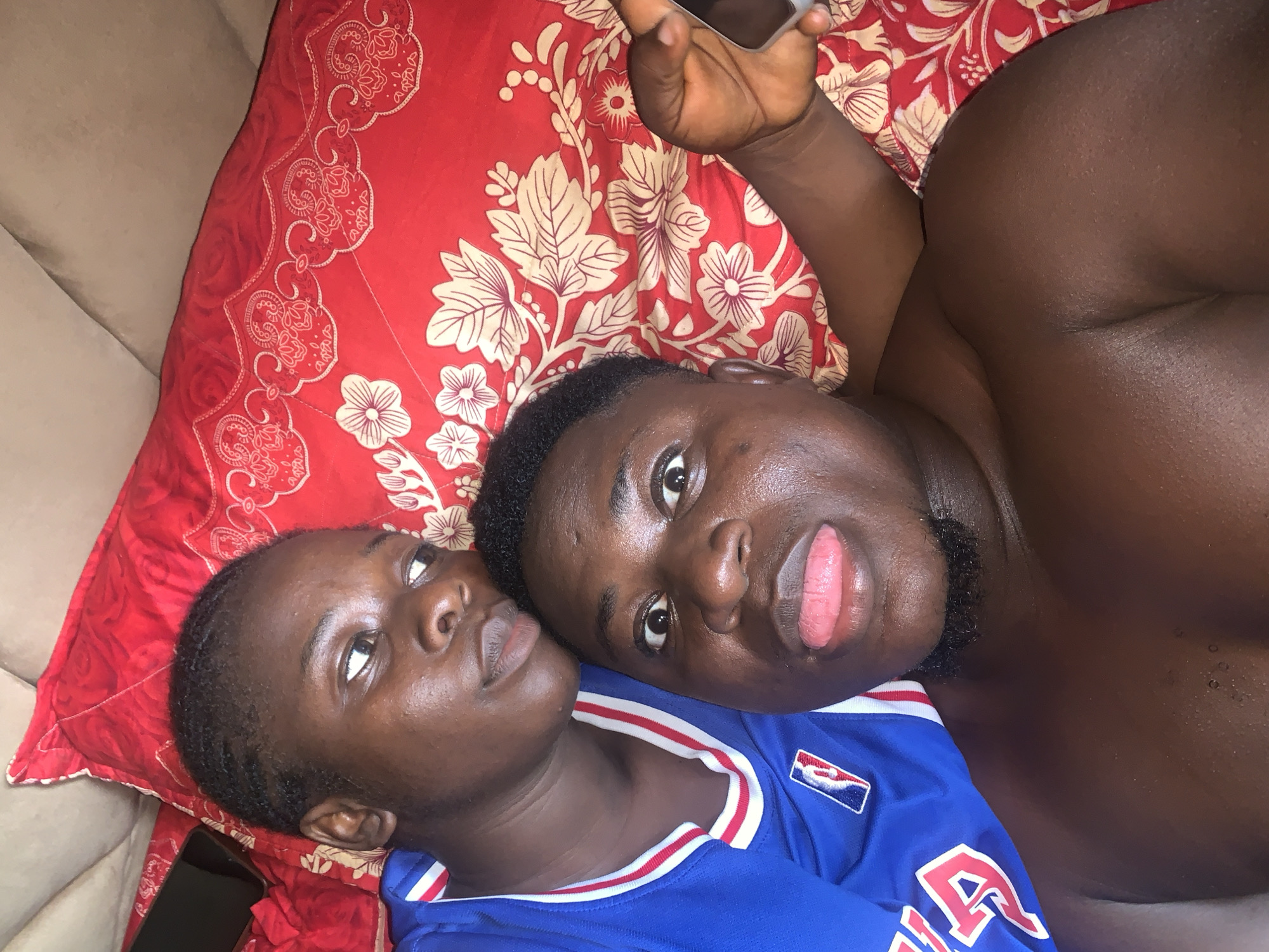 Two young Black individuals, a male and a female, are lying on a red bedspread with a floral pattern. The female, positioned behind and to the left of the male, wears a blue jersey with white and red trim and an NBA logo. Her head is turned to the left, and she looks away from the camera. The male, in the foreground and center, is shirtless and has a playful expression, with his tongue sticking out. He is holding a mobile phone in his right hand, extended towards the right side of the frame. They appear to be in an indoor setting, possibly a bedroom, in Akwanga, Nigeria. The lighting suggests it might be daytime or a well-lit indoor space.