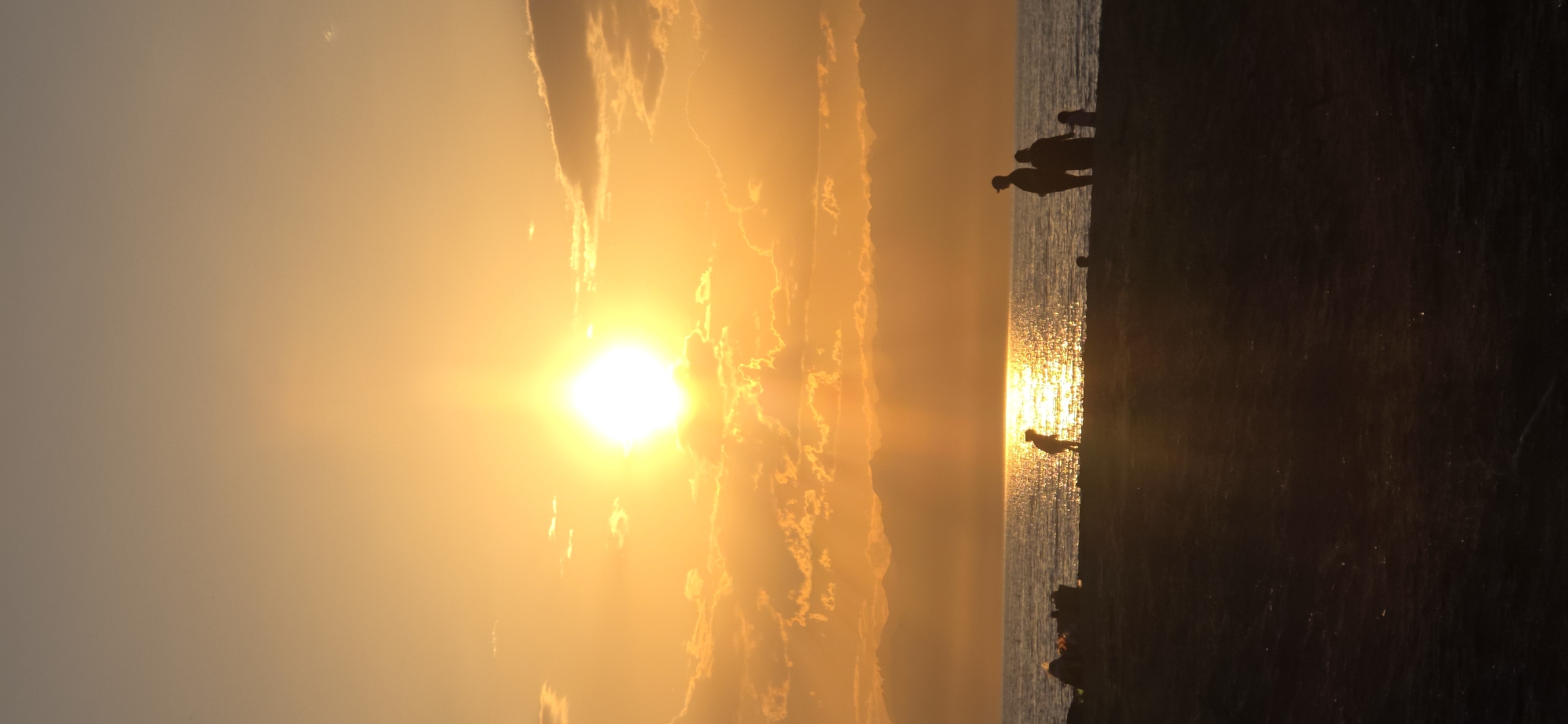 The image depicts a sunset over the Issyk-Kul lake near the village of Boz Beshik, Kyrgyzstan. The sun is a bright, intense orb in the sky, casting a warm, orange glow across the scene and creating a shimmering reflection on the water. The sky is filled with scattered clouds, some of which are illuminated by the setting sun, giving them a golden hue.
In the foreground, a silhouette of a beach or shore is visible, with a few figures of people scattered across it. To the left, near the water's edge, there appear to be some objects or debris. In the middle ground, a solitary figure, possibly a child, is silhouetted against the bright reflection of the sun on the water, bent over as if playing. Further to the right, another group of silhouettes, appearing to be a family with at least two adults and a child, stand facing the water and the sunset. The overall atmosphere is serene and tranquil, capturing the beauty of a sunset over a large body of water.