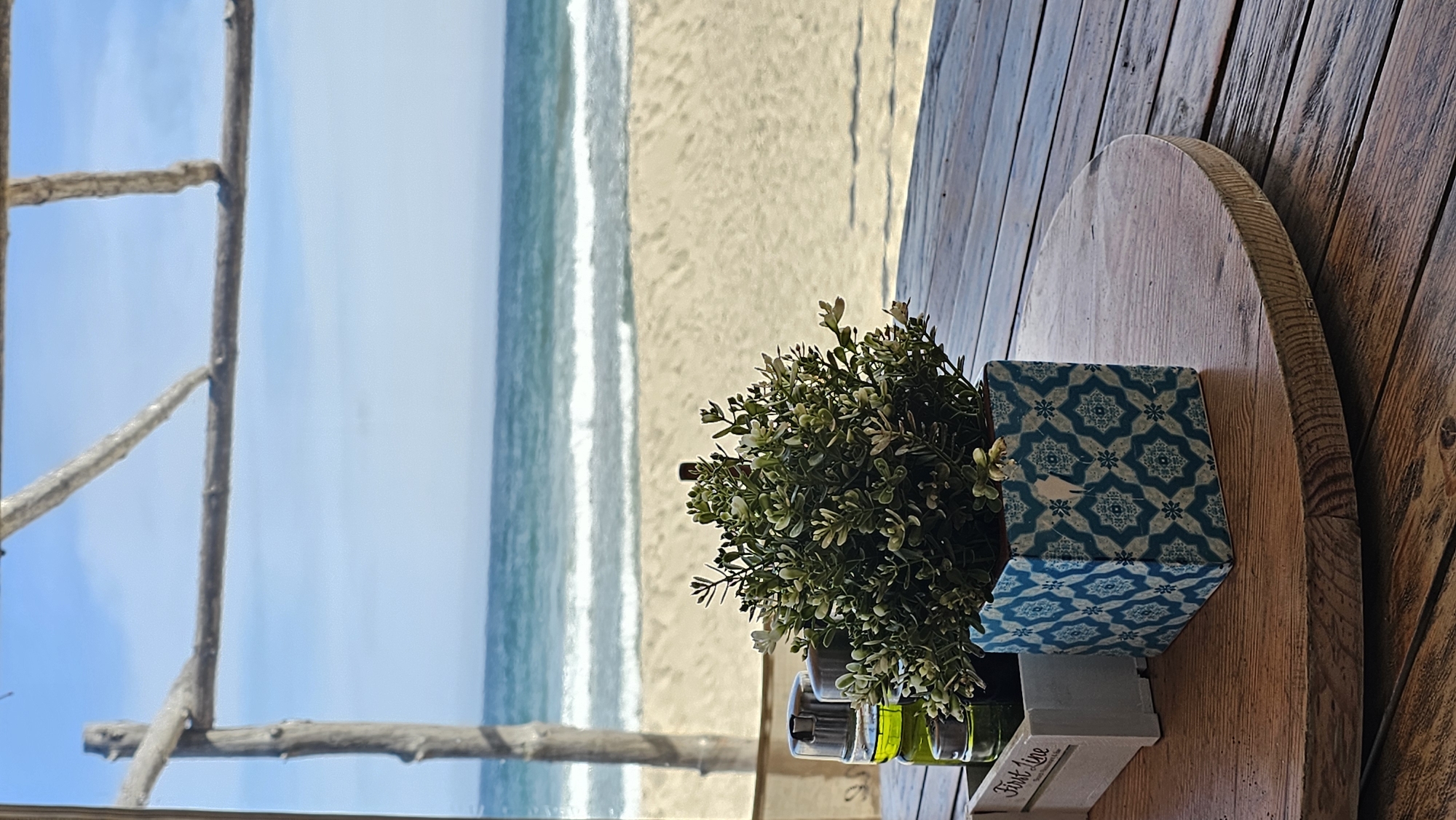 The image captures a serene beachfront view from what appears to be a cafe or restaurant in Kranevo, Bulgaria, likely taken during the day under a clear sky.
In the foreground, a rustic wooden table occupies the right side of the frame. On the table, a small potted artificial plant with light green foliage and tiny white flowers is nestled in a white wooden holder. Next to the plant, there's a small set of cruets, possibly for oil, vinegar, salt, and pepper, with two visible green-tinted bottles. A decorative blue and white patterned tile or coaster is also placed on the table. The white wooden holder prominently displays the text "First Line." The table itself is a light-colored wood, appearing to be part of a larger, darker planked wooden surface.
To the left, a natural, rustic window-like frame made of rough wooden branches or driftwood provides a portal to the outside scene. Beyond this frame, a wide expanse of light sandy beach stretches towards the calm, blue-green sea. Gentle white waves are seen breaking along the shoreline. The sea extends into the horizon under a clear, bright blue sky, creating a peaceful and inviting atmosphere.
The scene suggests a relaxing dining or lounging experience by the sea, emphasizing tranquility and natural beauty.