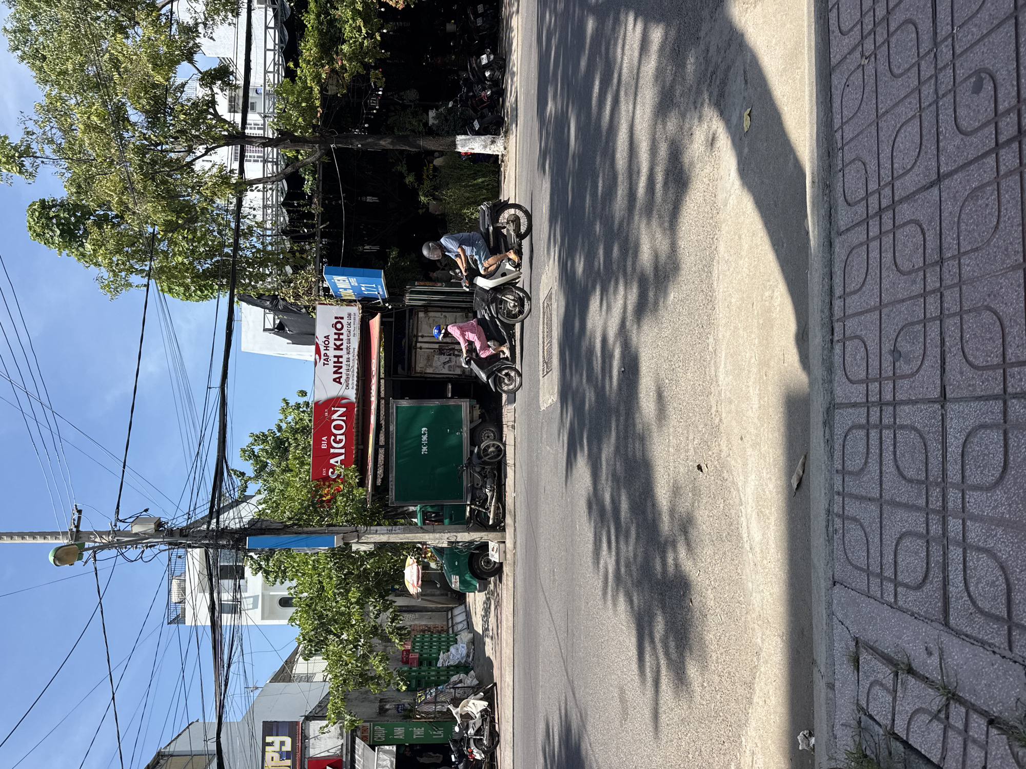 The image shows a street scene in Bac Nha Trang Ward, Vietnam. The foreground is dominated by a sidewalk made of grey tiles with a patterned design. Beyond the sidewalk is a paved road with faint shadows cast by overhead trees.
In the midground, two people are riding on separate motorbikes, traveling from left to right. The person on the left is wearing a pink outfit and a blue helmet, riding a dark-colored motorbike. The person on the right is wearing a blue shirt and shorts, and a clear helmet, riding a lighter-colored motorbike.
To the left of the road, there is a green delivery truck with the license plate "79C-196.79". Adjacent to the truck is a building with a red sign that reads "BIA SAIGON" and another sign that says "TẠP HÓA ANH KHỐI". In front of this building, there are stacks of green crates and some white bags.
Further back, there are more buildings, trees, and a complex network of electrical wires suspended from poles. The sky is clear and blue. The overall scene depicts a typical street environment in a Vietnamese city on a sunny day.