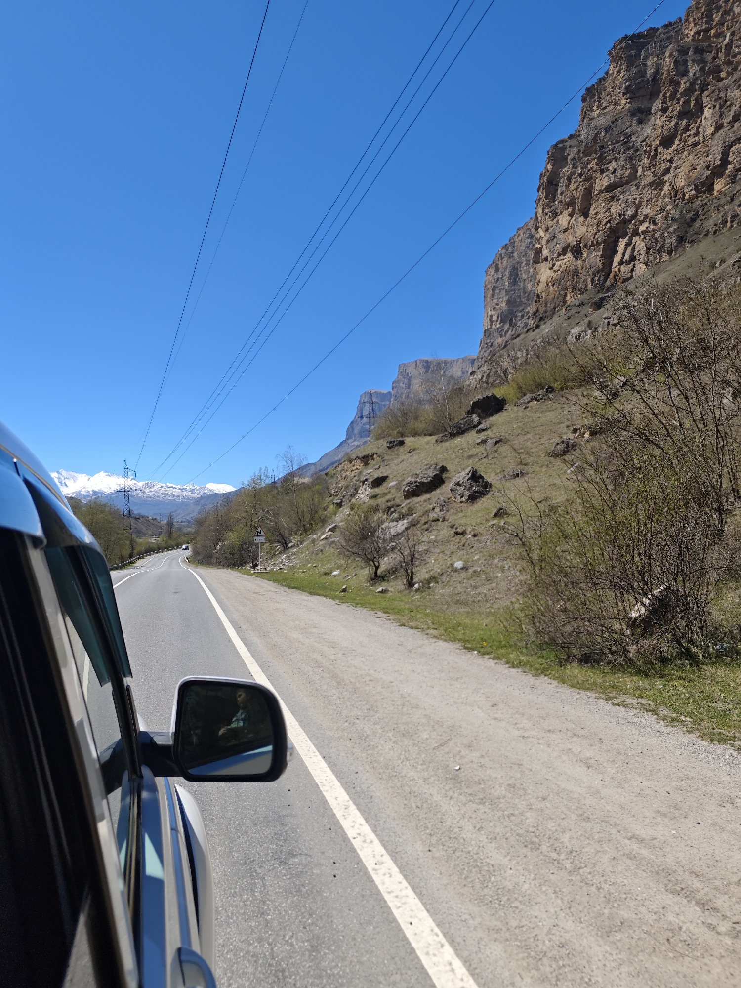 A daytime eye-level shot from inside a vehicle, showing a road in Bedyk, Russia. The road is asphalt, with a white line marking the right edge. To the right of the road is a gravel shoulder and a steep, rocky hillside. Bare trees and sparse vegetation cover the slope. A large, vertical rock face dominates the upper right. To the left of the road, there is a glimpse of a black car interior and side mirror reflecting a figure inside. In the distance, snow-capped mountains can be seen under a clear blue sky, crossed by power lines. The overall scene depicts a drive through a mountainous region on a sunny day.