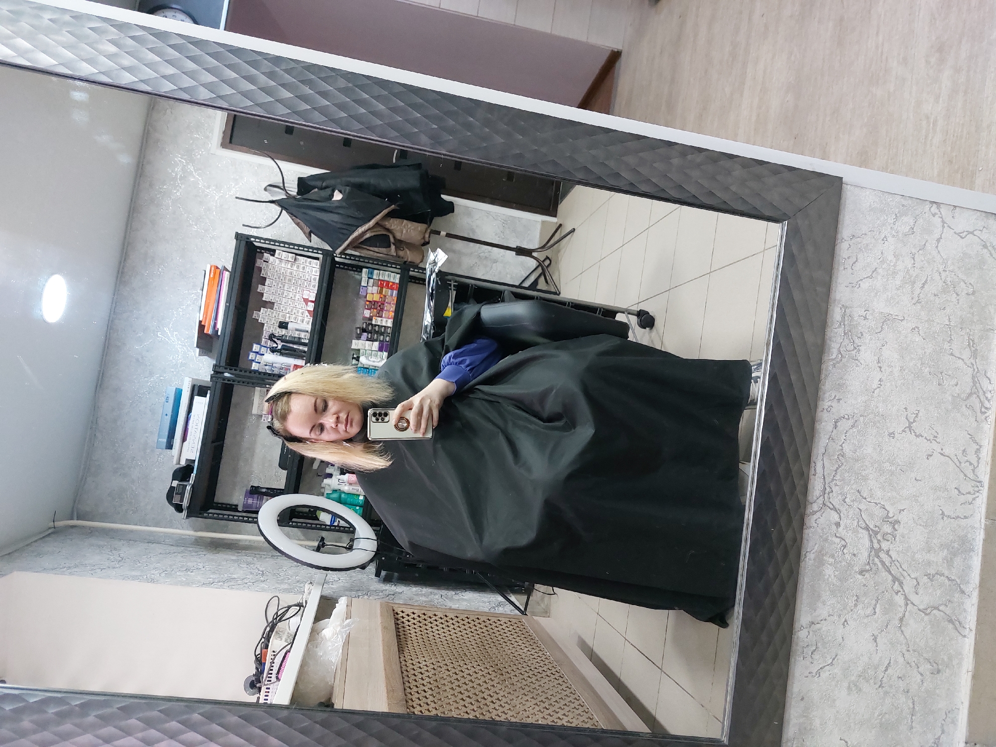 This image captures a mirror selfie taken by a woman in a hair salon or beauty studio in Chaikovsky, Russia. The photograph is rotated 90 degrees clockwise. The subject is a young woman with blonde hair, seen from the chest up, seated in what appears to be a black salon chair. She is wearing a blue top covered by a black styling cape, typically used during hair treatments. She holds a smartphone in her right hand, with its camera module visible on the back, suggesting she is taking the picture. Her eyes appear to be closed or cast downwards, giving a relaxed impression.
The reflection in the large mirror, which features a dark, geometrically patterned frame, shows the salon interior. Behind the woman, a black metal shelving unit is filled with numerous hair products, including various small boxes that likely contain hair dyes or treatments, along with books and folders. A white circular ring light on a stand is visible to her left, indicating professional lighting. The background also shows a light gray wall with a subtle marbled texture, and a light-colored tiled floor. A wooden radiator cover with a criss-cross pattern is visible in the lower part of the reflection, and a dark brown door or opening can be seen in the reflected background. The ceiling, reflected in the top portion of the mirror, has square-patterned tiles. The scene depicts a moment during a hair service, possibly a break, as the woman takes a selfie. No specific legible text is visible on any of the products or items.