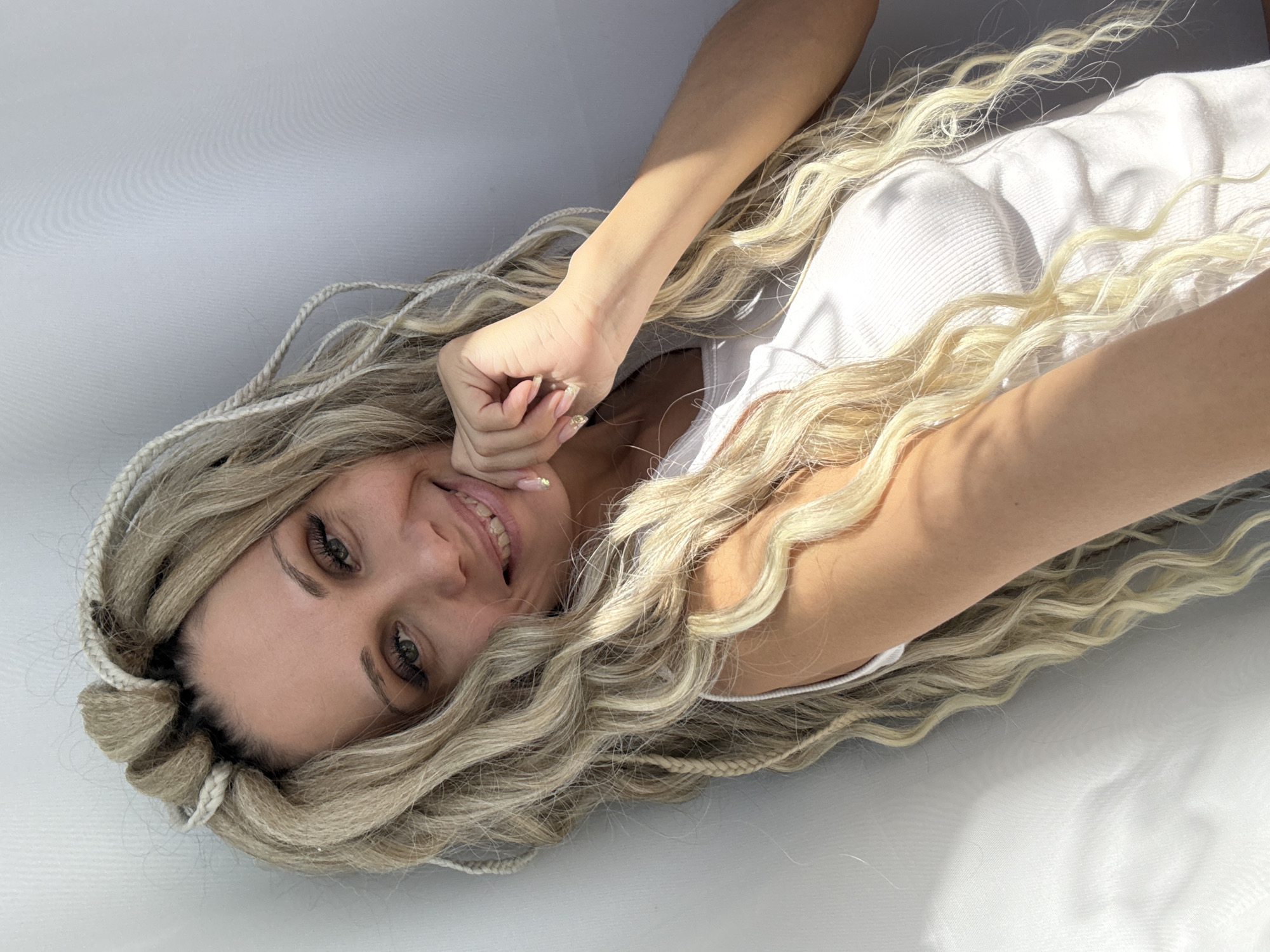 A woman with blonde hair poses in a white sleeveless shirt against a white surface. She is smiling and touching her face with her left hand. The location is not specified and there is no mention of Rusanovo, Russia.