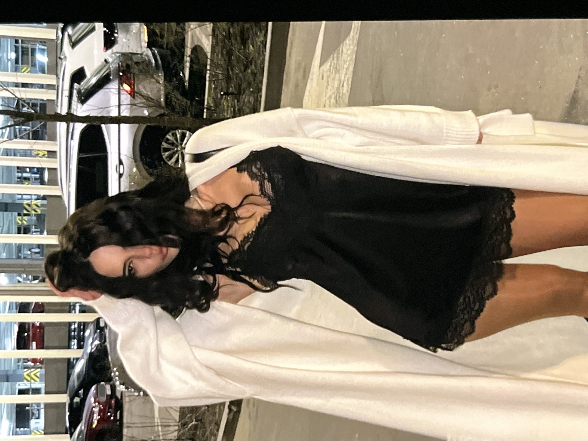 A woman with long, dark, wavy hair, wearing a black lace-trimmed slip dress and a white cardigan, stands on a paved surface outdoors at night. She is looking at the camera. In the background, there are cars parked in what appears to be a multi-story parking garage. The lighting suggests artificial illumination from the garage and possibly streetlights.