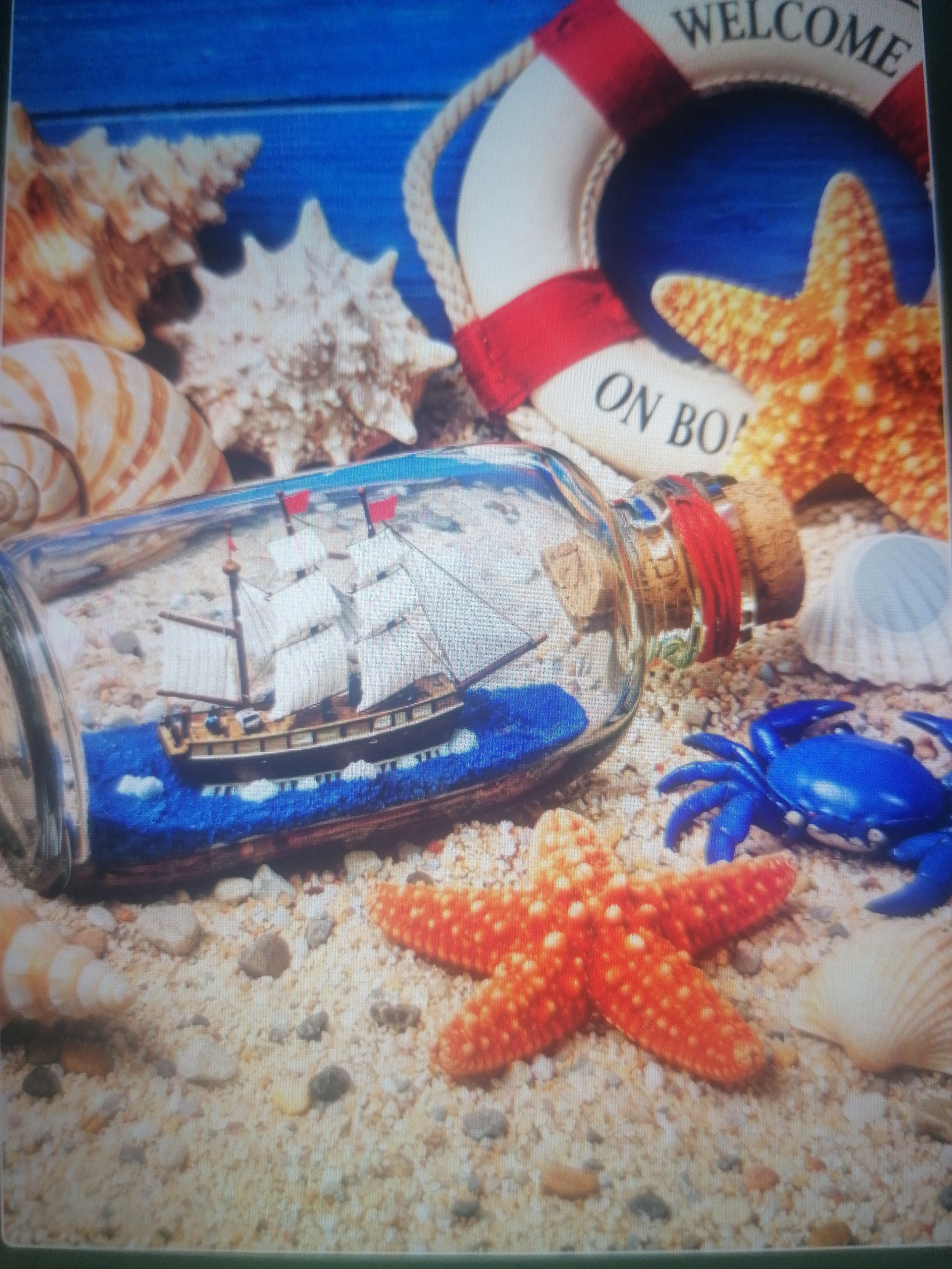 The image is a still life arrangement of nautical-themed objects on a sandy surface, with a blue wooden background. In the foreground, a clear glass bottle lies on its side, containing a miniature sailing ship with white sails and red flags. The bottle is stoppered with a cork wrapped in red ribbon. To the right of the bottle, a bright blue plastic crab is positioned. In the background, a life preserver with red and white stripes and the words "WELCOME ON BO" is visible. Various seashells, including a large orange starfish and a light-colored spiral shell, are scattered around the scene. The overall impression is a decorative tabletop display. There is no indication of a city or specific location.