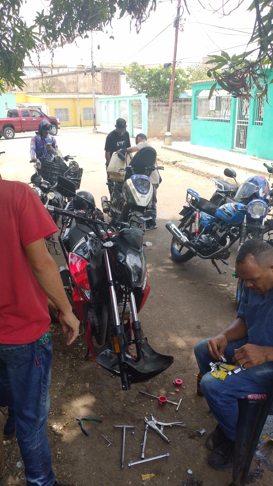 The image shows a street scene in Tinaquillo, Venezuela, where several people are gathered around motorcycles. One man is seated, working on a part of a motorcycle, with tools scattered on the ground nearby. Another man is standing by a red motorcycle, while others are attending to different motorcycles in the background. A red pickup truck is parked on the street in the distance. The surrounding area features houses with teal and yellow exteriors.