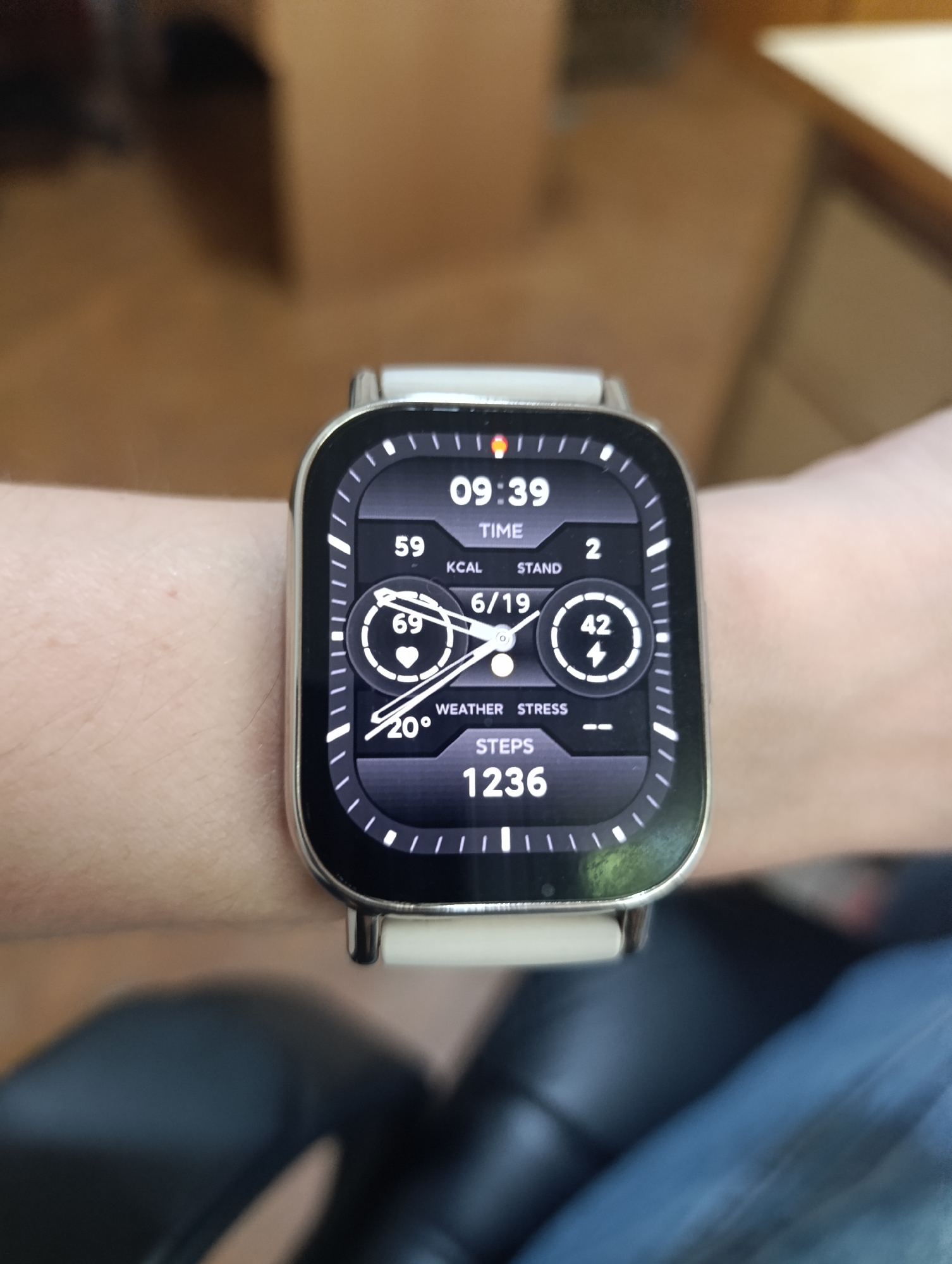 The image shows a close-up of a smartwatch on a person's wrist. The watch face displays the time as 09:39, along with various metrics including 59 KCAL, 2 STAND, 69 (heart rate), 6/19, 42 (lightning bolt icon), 20° (temperature), and 1236 STEPS. The background is blurred and appears to be an indoor setting with wooden furniture and dark seating. The scene is not located in a city.