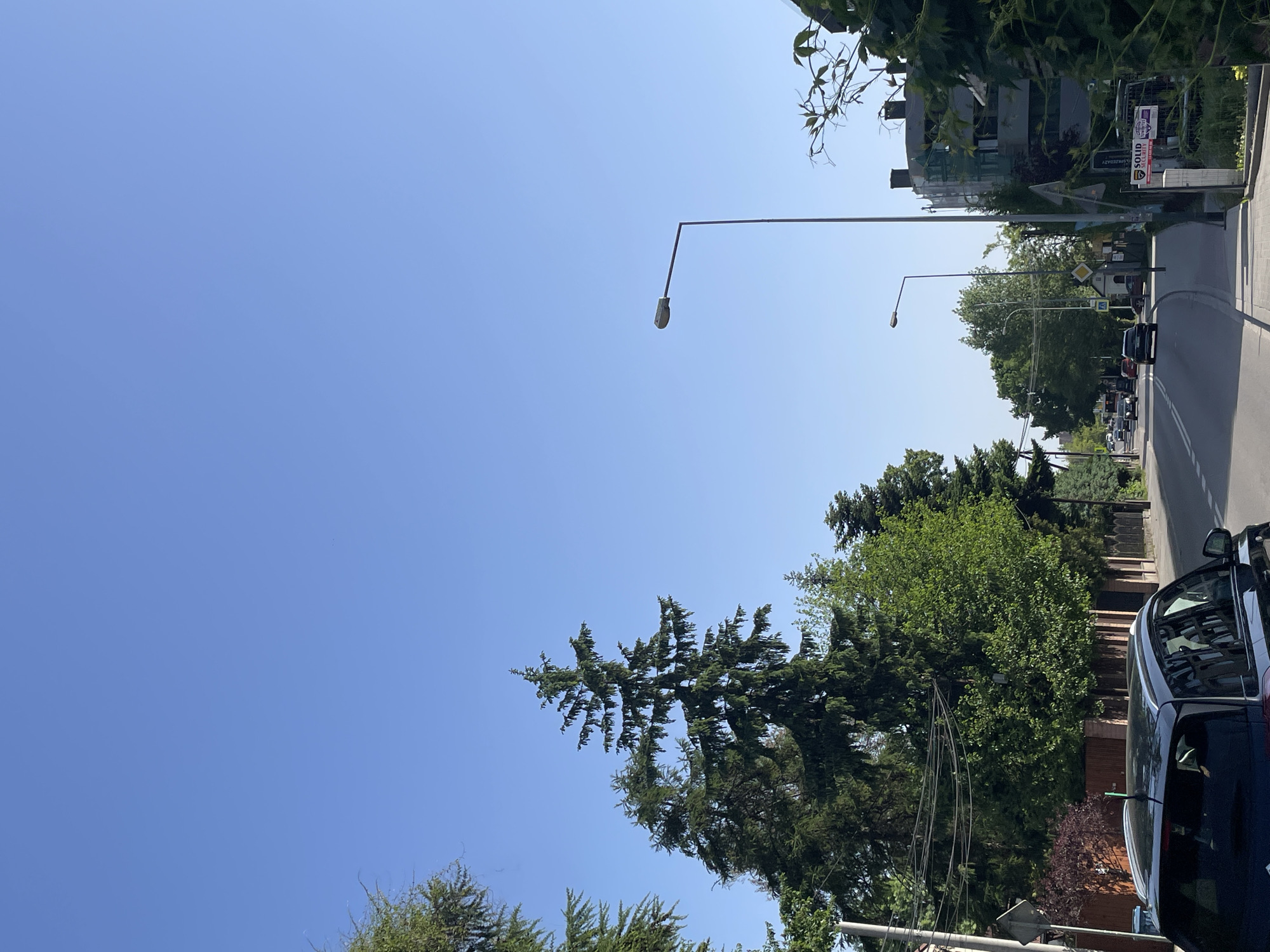 The image depicts a street scene in an urban location. The sky is clear blue, taking up a significant portion of the frame. Streetlights are visible extending over the road. Trees line the street, and a row of buildings can be seen in the background. A row of cars can be seen parked or driving along the road. A blue vehicle is parked on the right of the frame. A street sign is visible on the right.