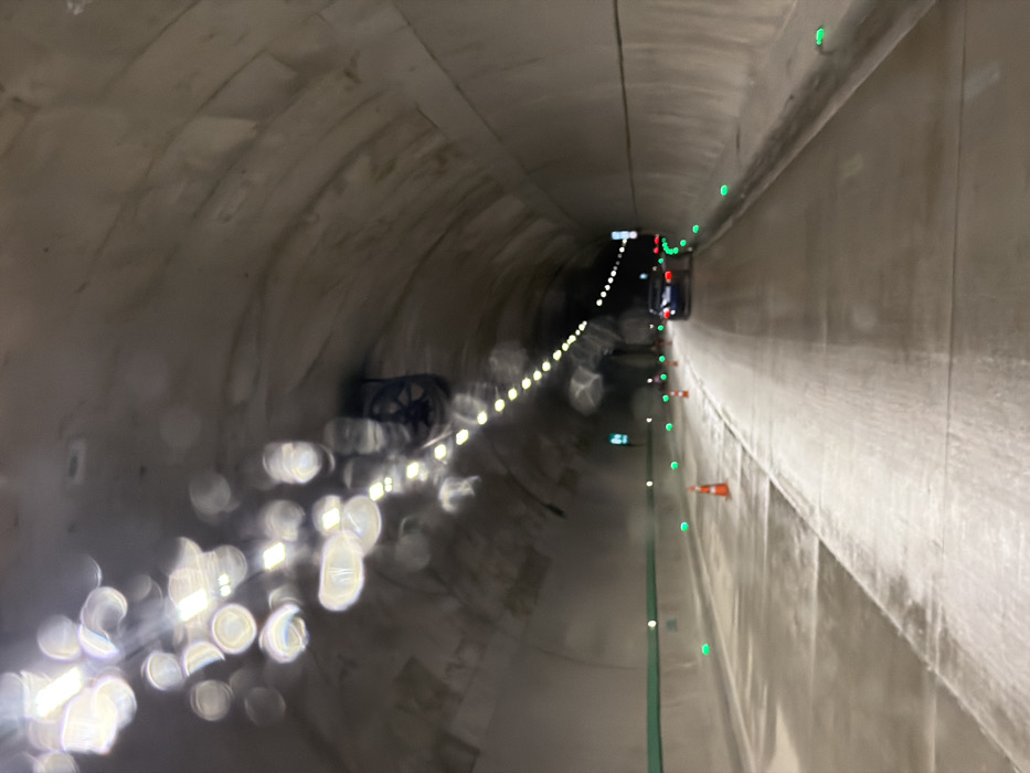 This image depicts the interior of a road tunnel located in Terjola, Georgia. The tunnel features an arched concrete ceiling and walls, with a continuous line of recessed, bright lights illuminating the roadway. A large, dark ventilation fan is mounted on the ceiling in the mid-ground. The road surface is concrete and appears damp, reflecting the overhead lights. The perspective is from inside a vehicle, looking forward into the tunnel.
The tunnel contains at least two lanes. The left lane is partially marked by a series of orange traffic cones placed along its length, indicating a potential work zone or partial closure. Green reflective markers or embedded lights are visible along both edges of the road. A dark-colored sedan, possibly blue or black, with illuminated red taillights, is driving ahead in the right lane, receding from the viewer. No individuals are visible. The foreground shows multiple circular and elongated light refractions, consistent with water droplets on a windshield or camera lens.