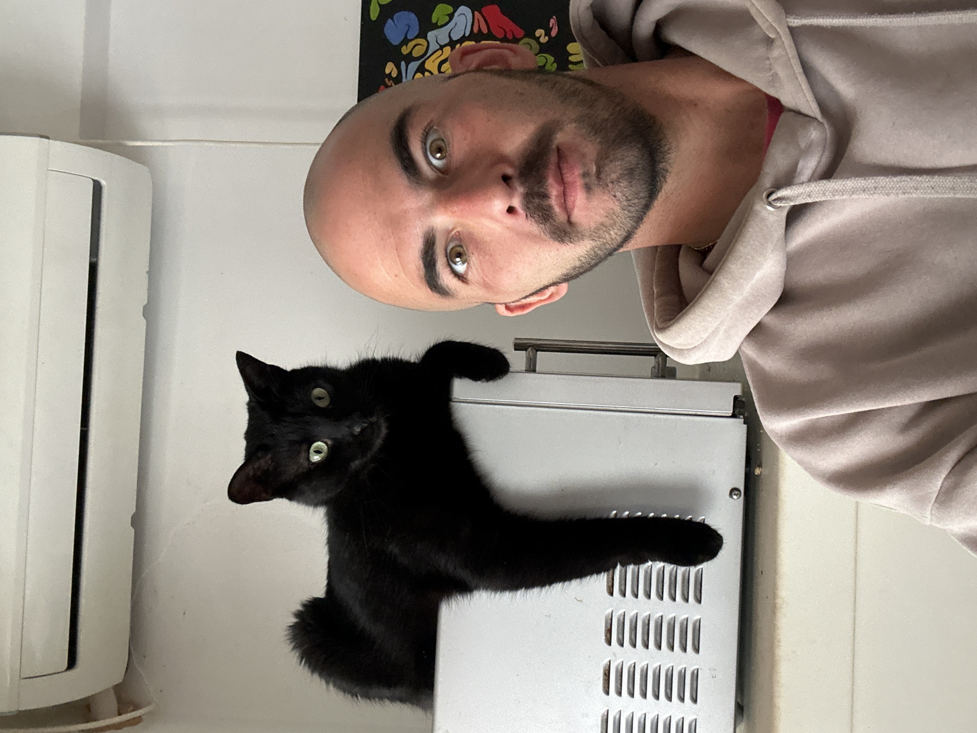 A rotated selfie shows a bald man with a beard wearing a hoodie posing with a black cat. The cat is perched on top of a silver metal appliance next to an air conditioning unit. The location is possibly Budva, Montenegro, given the user's profile.
