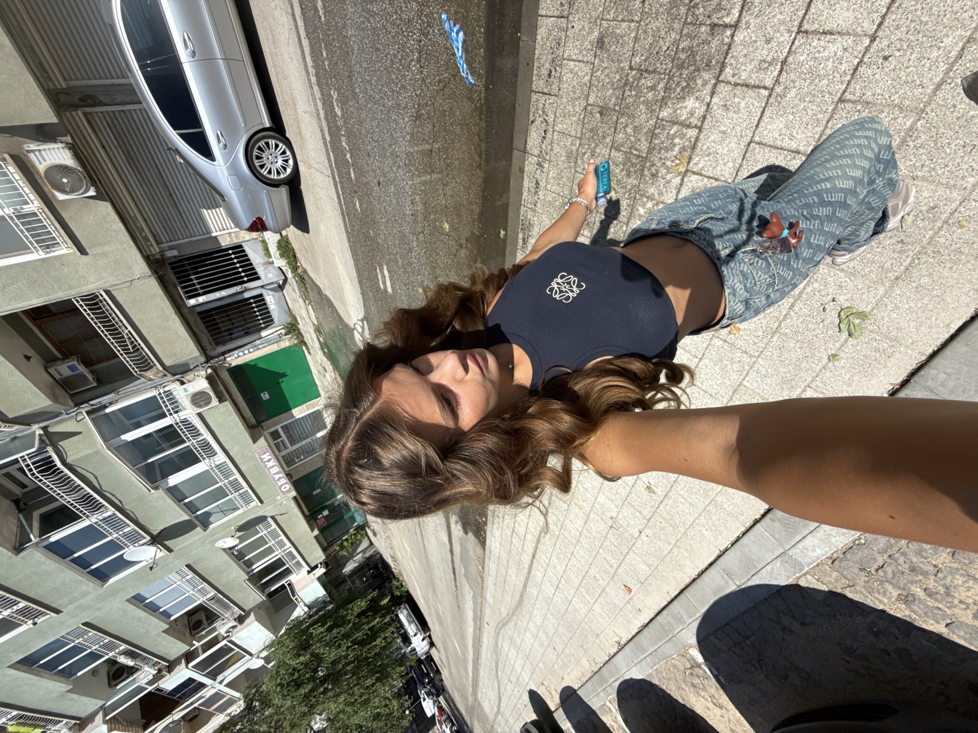 A young woman with long, wavy brown hair takes a selfie while walking on a sidewalk in Plovdiv, Bulgaria. She is wearing a dark crop top with a logo and patterned jeans. She holds a turquoise object in her right hand. The background shows a street with parked cars and a multi-story building with balconies and air conditioning units. The building's facade is a muted green. A silver car is parked on the street. The scene appears to be a sunny day.