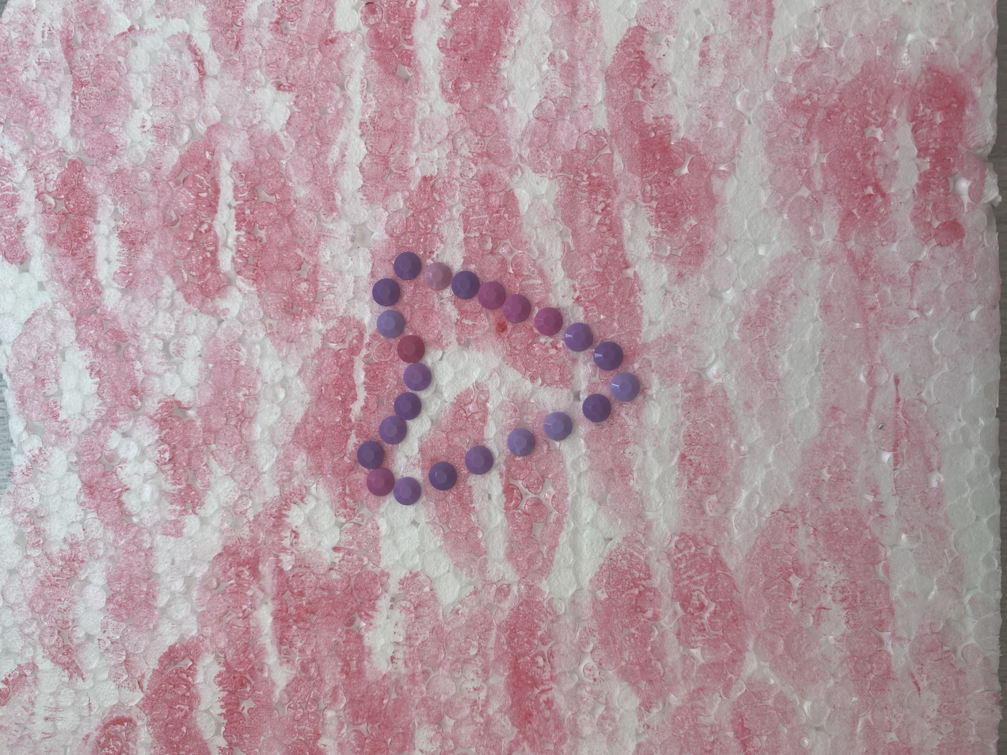 The image displays a close-up, overhead view of a piece of white textured material, possibly styrofoam, adorned with pink imprints resembling lipstick kisses arranged in horizontal stripes. In the center of the frame, a heart shape is formed by small, round, purple and pink gems. The texture of the material is irregular, with numerous circular indentations. The lighting is even, suggesting an indoor setting. There is no discernible geographical context, and therefore, no specific location in Blagoevgrad, Bulgaria, can be determined. The image depicts a static arrangement of objects with no discernible action or interaction.