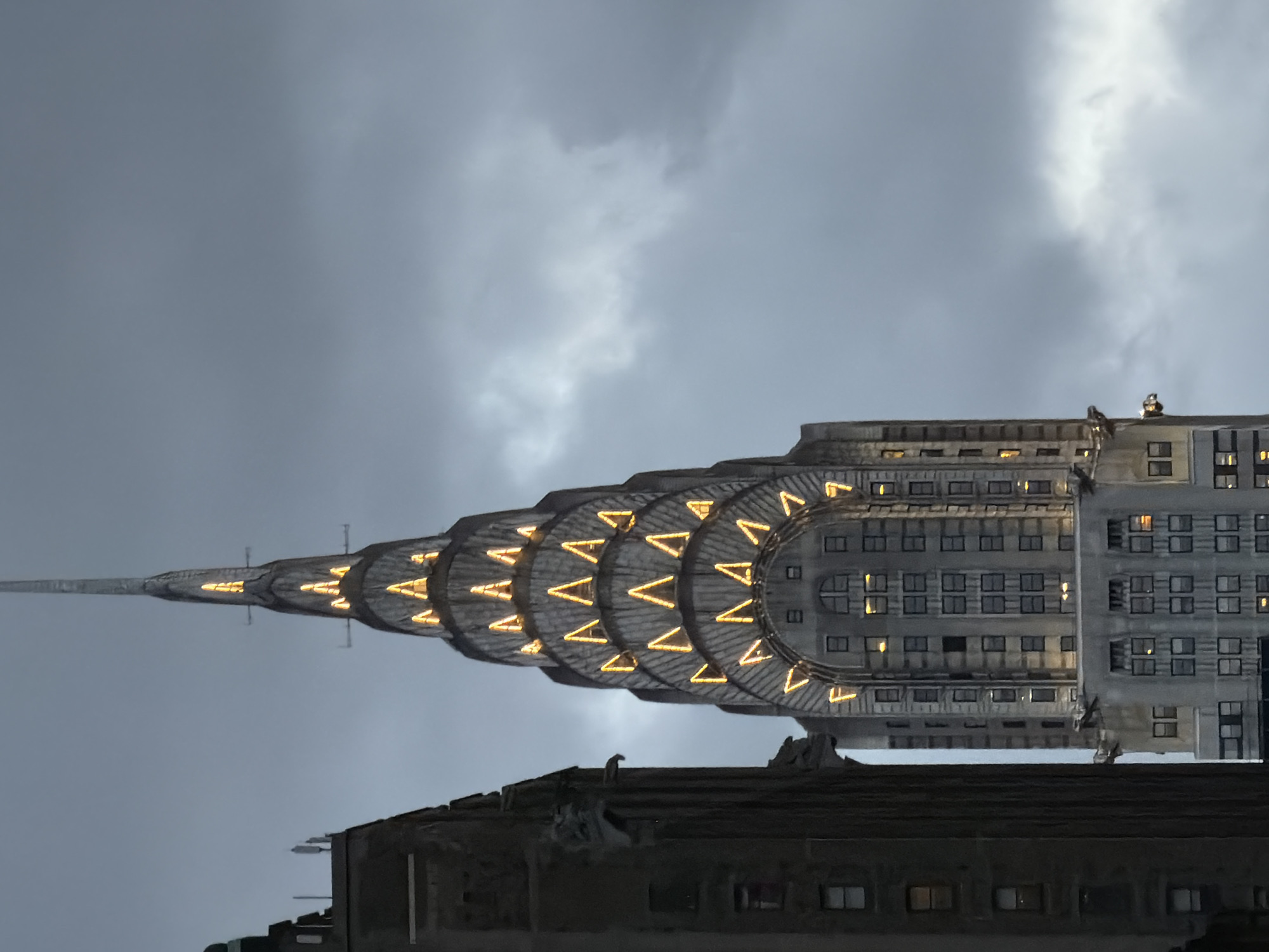 The image depicts the Chrysler Building in New York City, United States of America. The building's distinctive spire and illuminated ornamental details are visible against a backdrop of an overcast sky. The angle is from below, looking up at the building's upper portion. In the foreground, the top of another building partially obscures the lower portion of the Chrysler Building. There is no indication of the date this photo was taken. There are two subjects at the top of the closest building.
