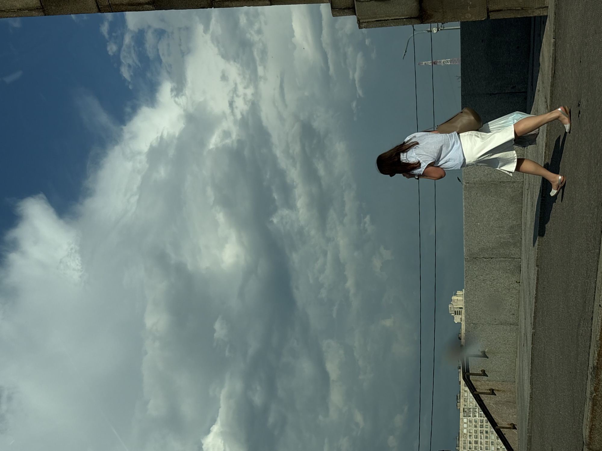 A woman with long brown hair, wearing a blue and white striped shirt and a white skirt, walks away from the camera along a paved path. She carries a brown bag over her shoulder and a white plastic bag in her hand. In the background, a large apartment building with many windows and a radio tower are visible under a cloudy sky. The scene appears to be a pedestrian area in the city of Saint Petersburg, Russia.