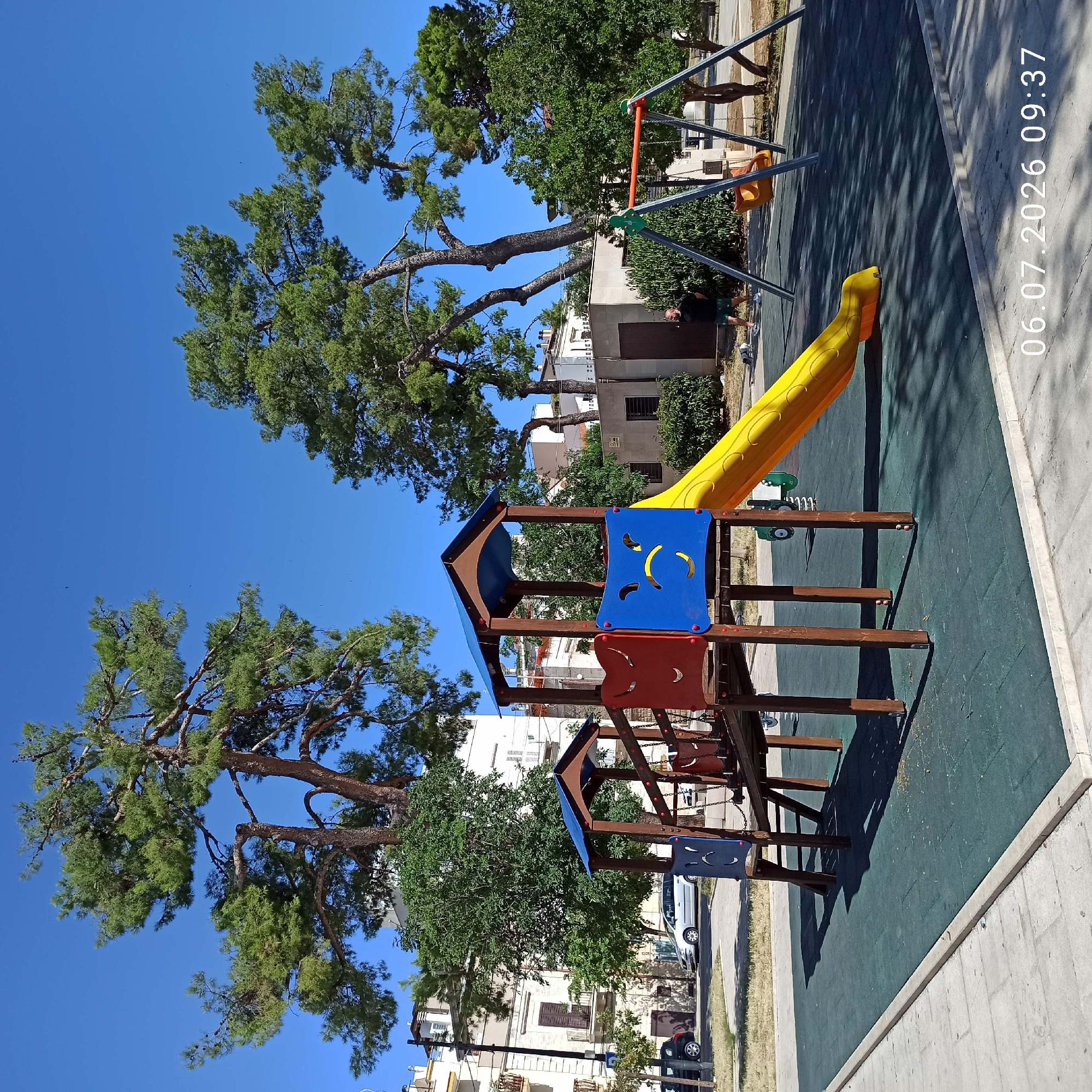 This is a full, eye-level shot of a children's playground in Ruvo di Puglia, Italy, on a sunny day. The playground features a wooden play structure with a blue roof, red and blue panels, and a yellow slide. To the right of the play structure, there is a swing set with two swings. The playground is situated on a dark green rubberized surface, bordered by a concrete path. In the background, there are large pine trees, lush green bushes, and buildings, suggesting an urban or suburban setting. A man wearing a black t-shirt and green shorts is standing near the swing set, accompanied by two dogs. Cars are parked along the street in the distance. The date and time stamp "06.07.2026 09:37" is visible in the lower right corner.
