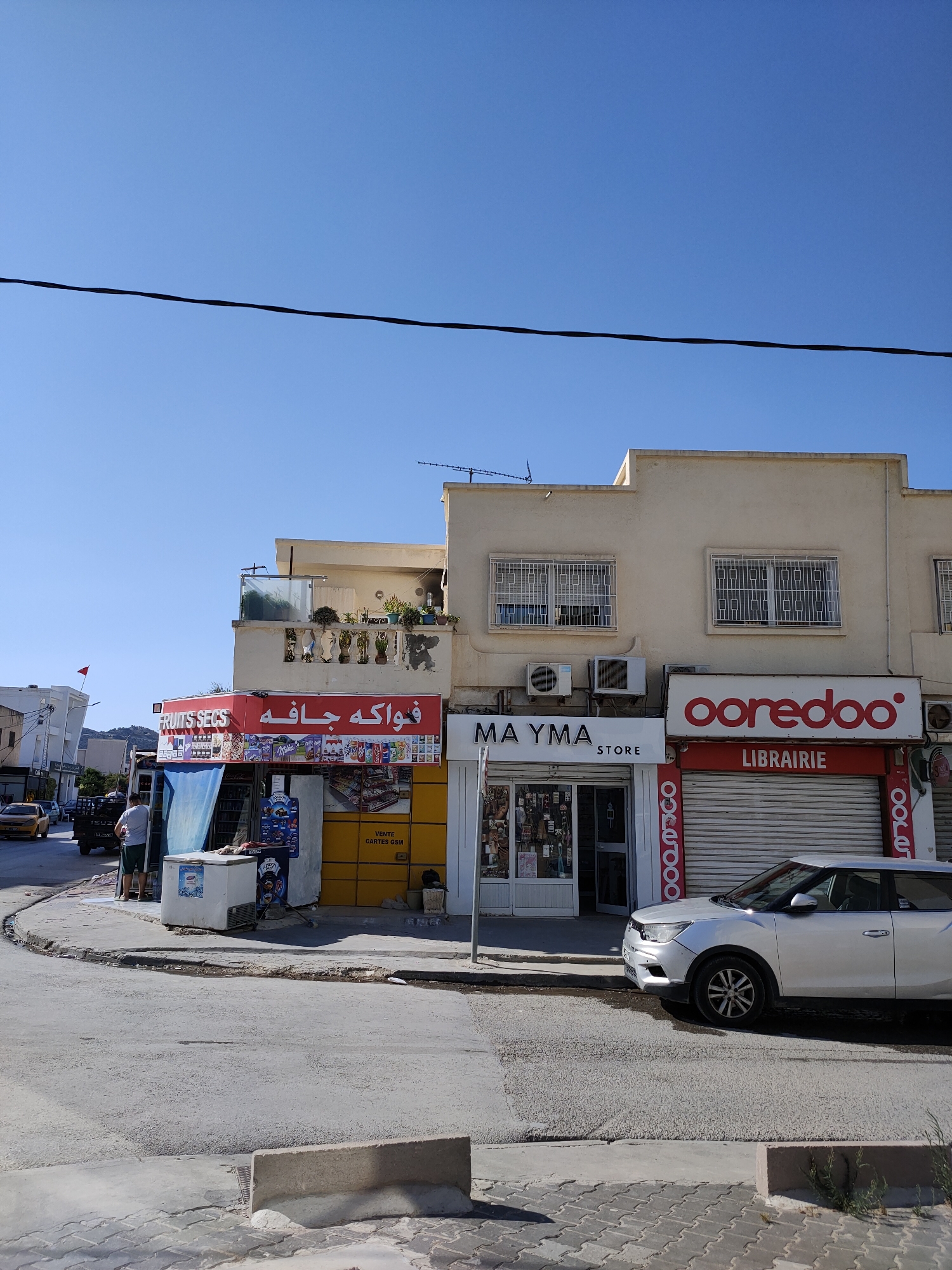In an urban street scene under a clear blue sky, the focal point is a row of commercial establishments. The building features a pale yellow facade with multiple stories and balconies adorned with potted plants. Prominently displayed are store signs: "FRUITS SECS" and Arabic text are above a shop displaying various products, including a blue tarp and an open freezer. To its right, a store labeled "MA YMA STORE" has a glass storefront showcasing merchandise. Further to the right, a bright red and white sign reads "ooredoo" above a rolling metal gate labeled "LIBRAIRIE."
In the foreground, a silver SUV is parked on the asphalt street, which shows signs of wear and some water accumulation. The street is bordered by a sidewalk paved with interlocking bricks. A concrete barrier is positioned near the bottom of the frame. In the background, a yellow car and a dark-colored vehicle are visible on the road, with other buildings and some greenery beyond. A person in a white t-shirt and shorts stands near the "FRUITS SECS" shop, looking towards the store. A small red flag is visible on a pole on a building to the left. The overall environment suggests a commercial district in a town.