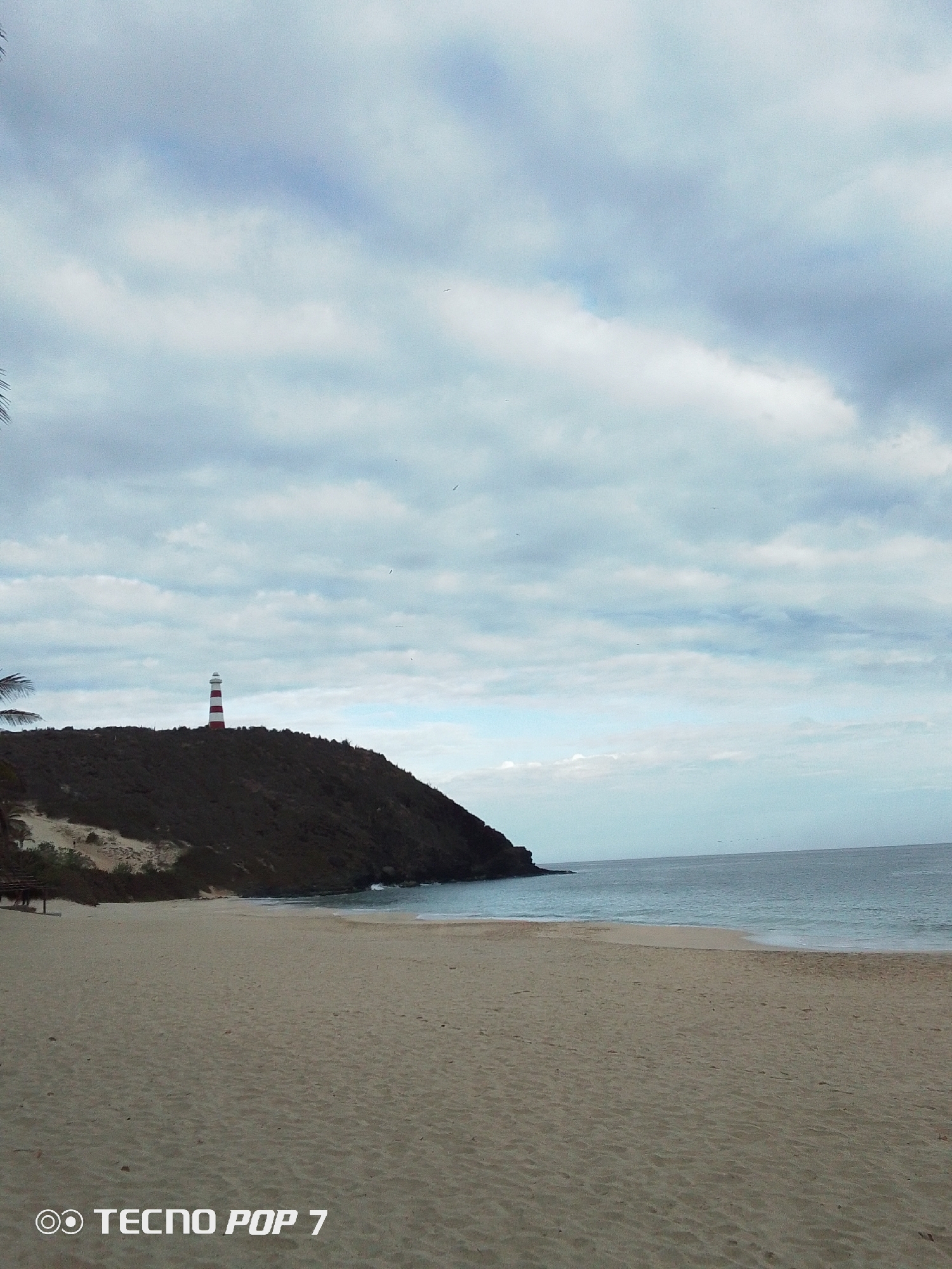 The image depicts a beach scene in Pedro González, Venezuela, dominated by a sandy beach in the foreground. The beach leads to calm, light blue water meeting the shoreline. In the midground, a dark, vegetation-covered hill rises, topped by a red and white striped lighthouse. The sky is overcast with a mix of light and dark clouds. A small, covered structure is visible on the left edge of the hill, near the beach. The words "TECNO POP 7" are visible in the lower left corner, indicating the device used to capture the image.