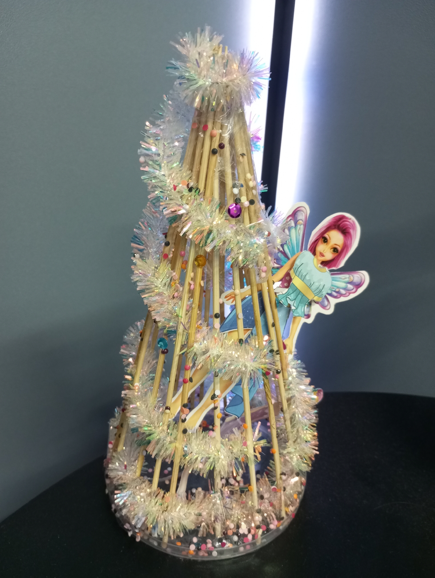 A decorated Christmas tree-shaped object is placed on a dark, reflective surface. The object appears to be handmade, with a frame made of wooden sticks, wrapped in tinsel and adorned with small, colorful beads. A cutout of a fairy with pink hair and blue wings is attached to one side of the tree. The background consists of a light gray wall and a bright, vertical light source. The exact location in the city of Pervomaysky, Russia is not evident from the image.