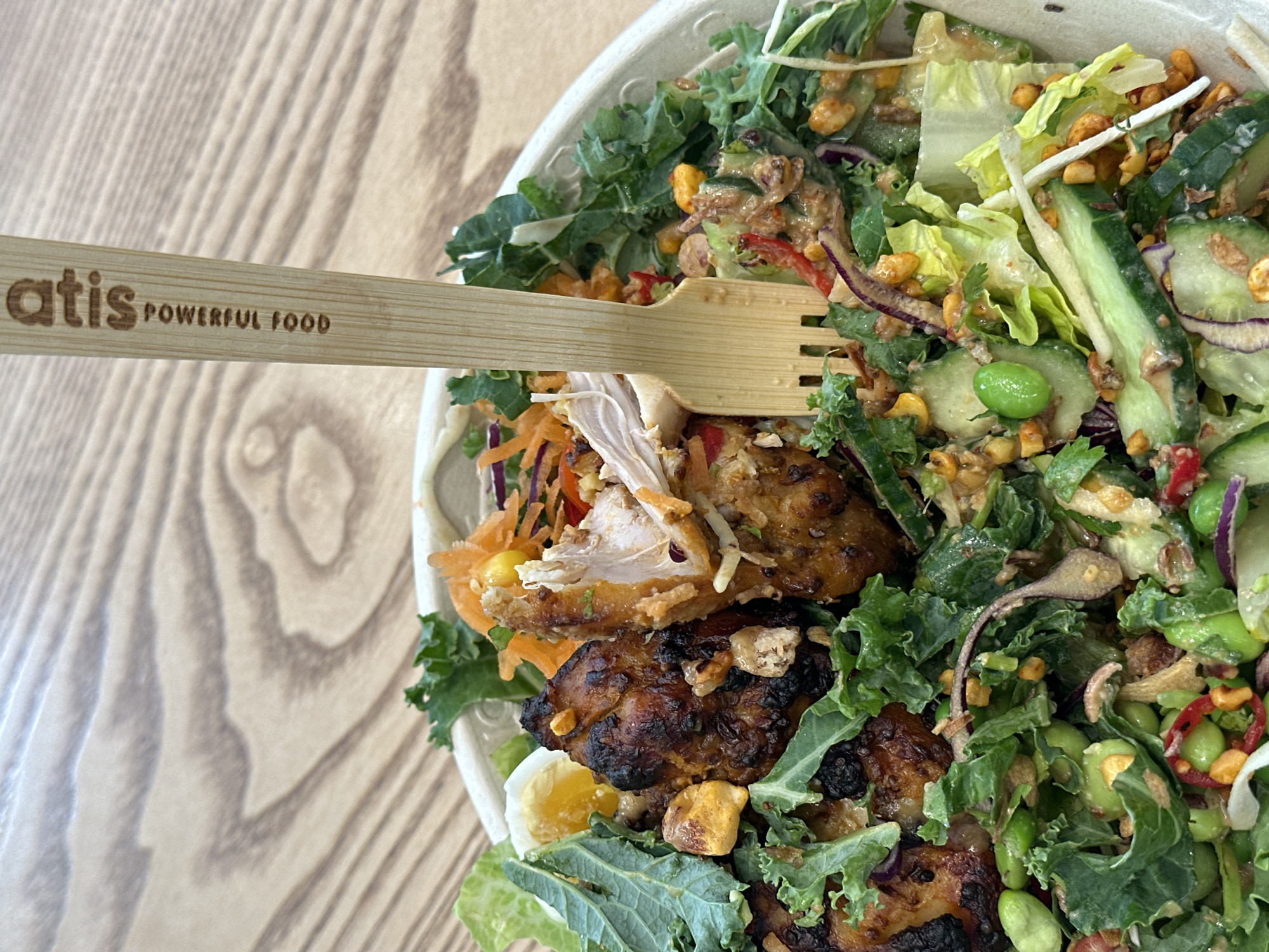 A high-angle shot displays a partially consumed salad in a white bowl, accompanied by a light-colored wooden fork. The salad contains kale, lettuce, cucumber, edamame, shredded carrots, red onion, and pieces of dark-browned chicken. A hard-boiled egg is visible near the bottom left. The fork has the words "atis POWERFUL FOOD" branded on it. The bowl sits on a light-colored wooden table. The Atis restaurant chain is located in London, United Kingdom.